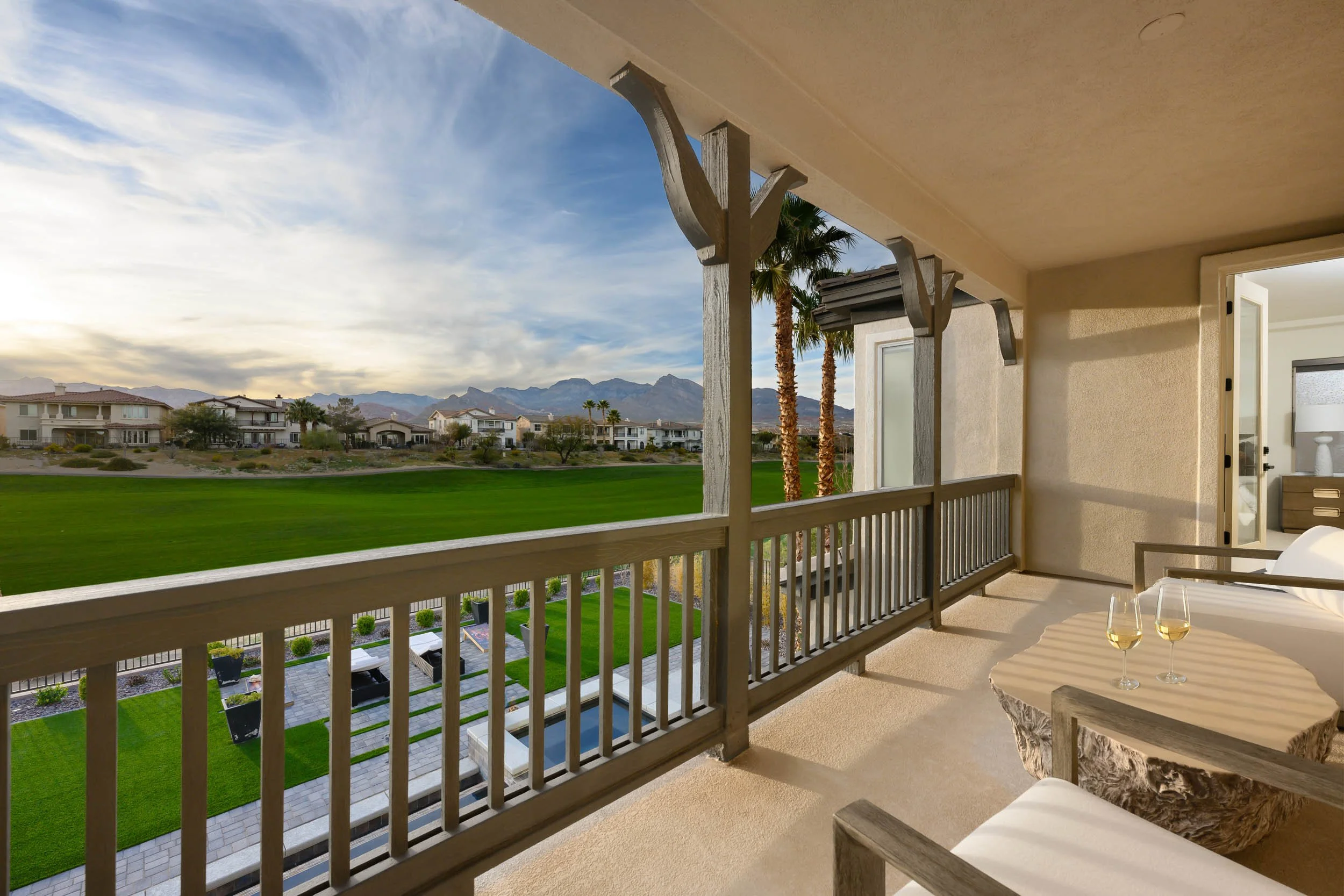 Balcony with two glasses of white wine on a wooden table, overlooking a golf course and residential neighborhood with mountain range in the distance.