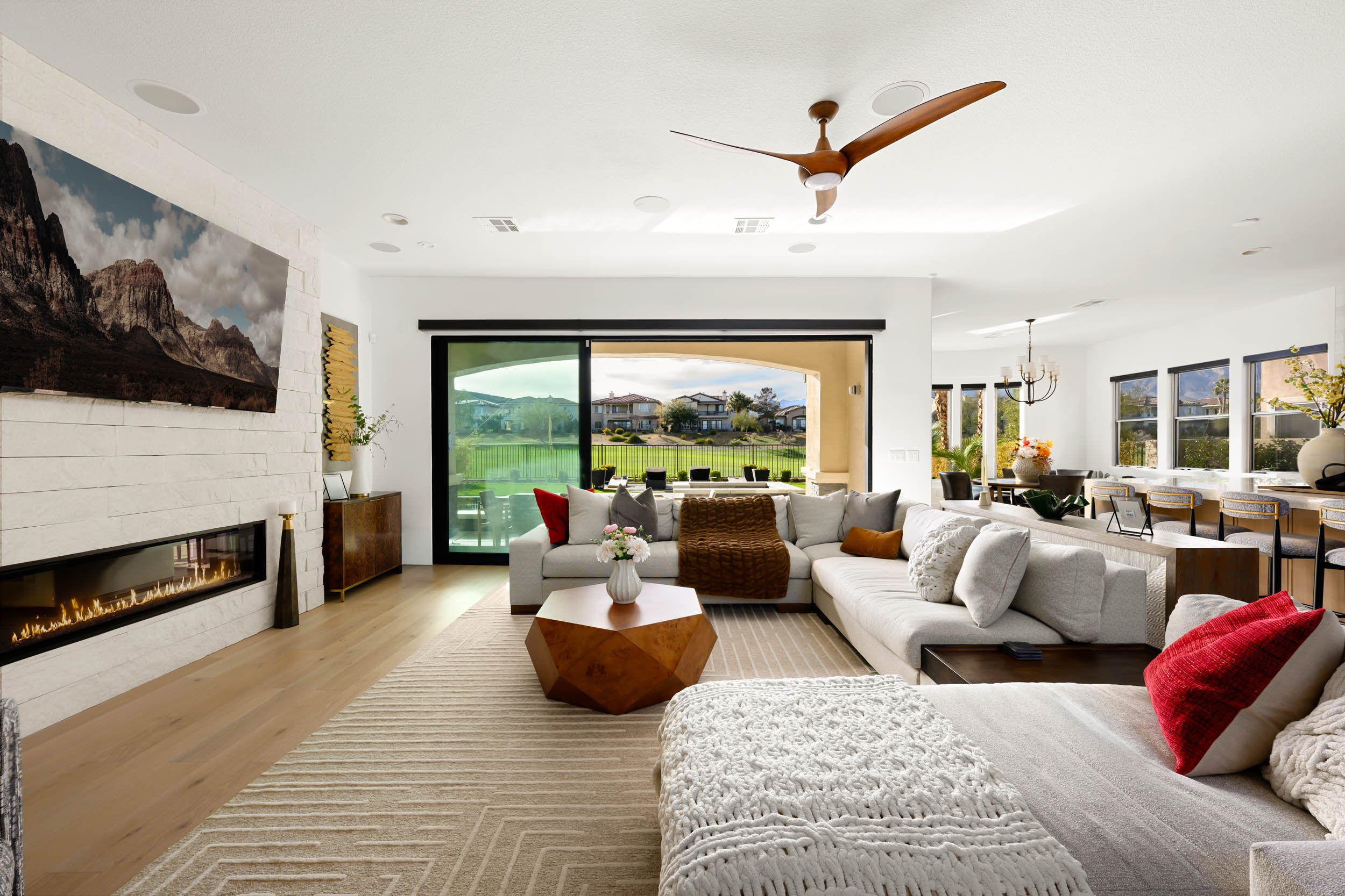 A spacious living room with a white sectional sofa, colorful pillows, a geometric wooden coffee table, and a modern fireplace with a mountain picture above it. Large windows and sliding glass doors offer a view of a green backyard and neighboring hou