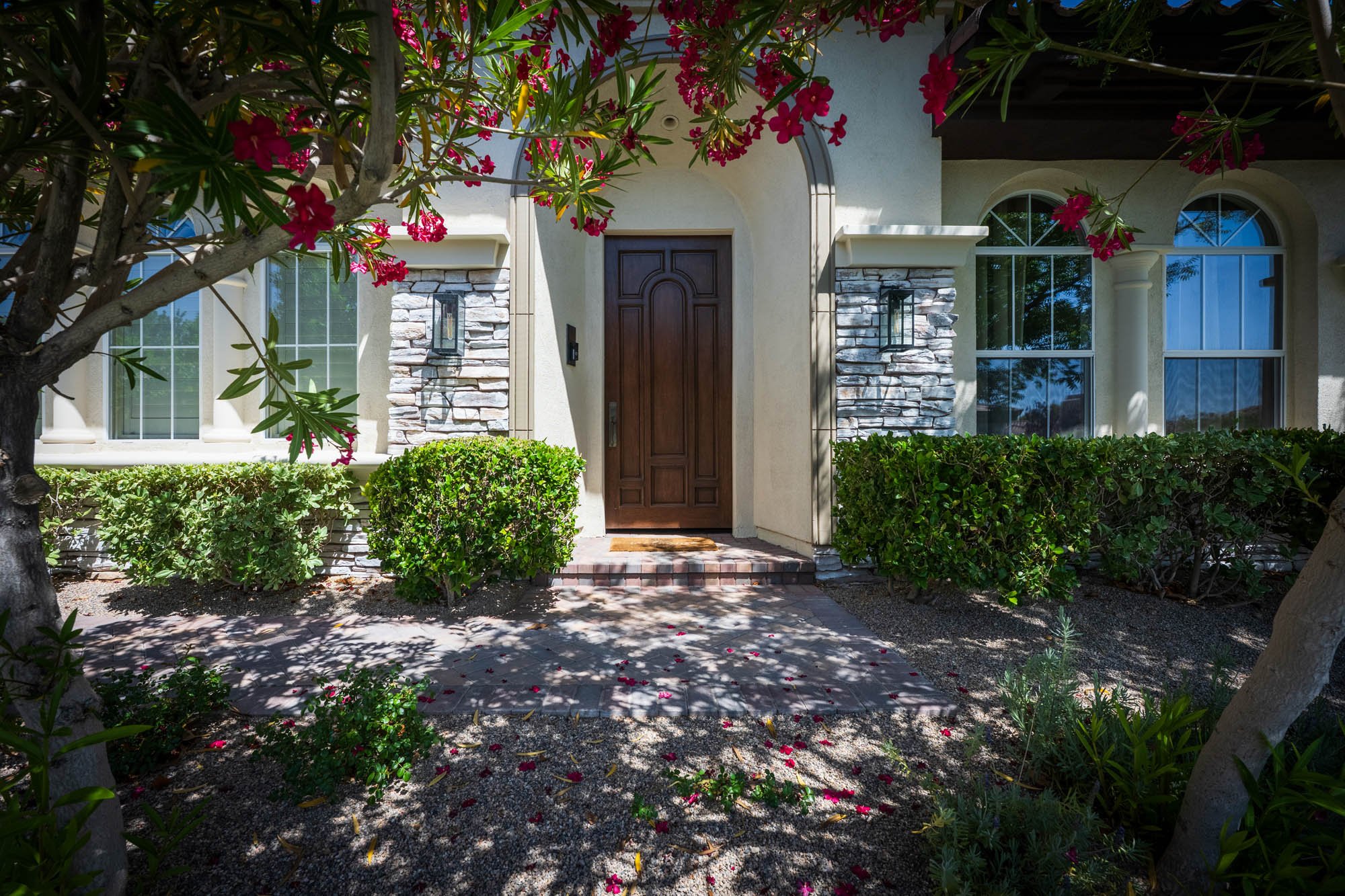 Front view of a house with a wooden door, stone accents, and large windows, surrounded by bushes, trees, and pink flowers in a landscaped yard.
