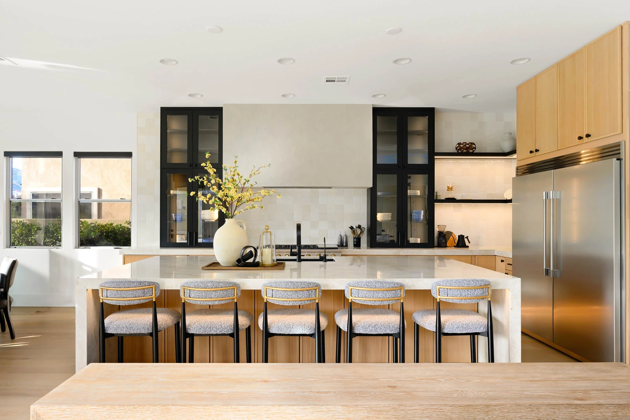 Modern kitchen with a large marble island, black and wood cabinetry, and stainless steel appliances, decorated with a vase of yellow flowers.
