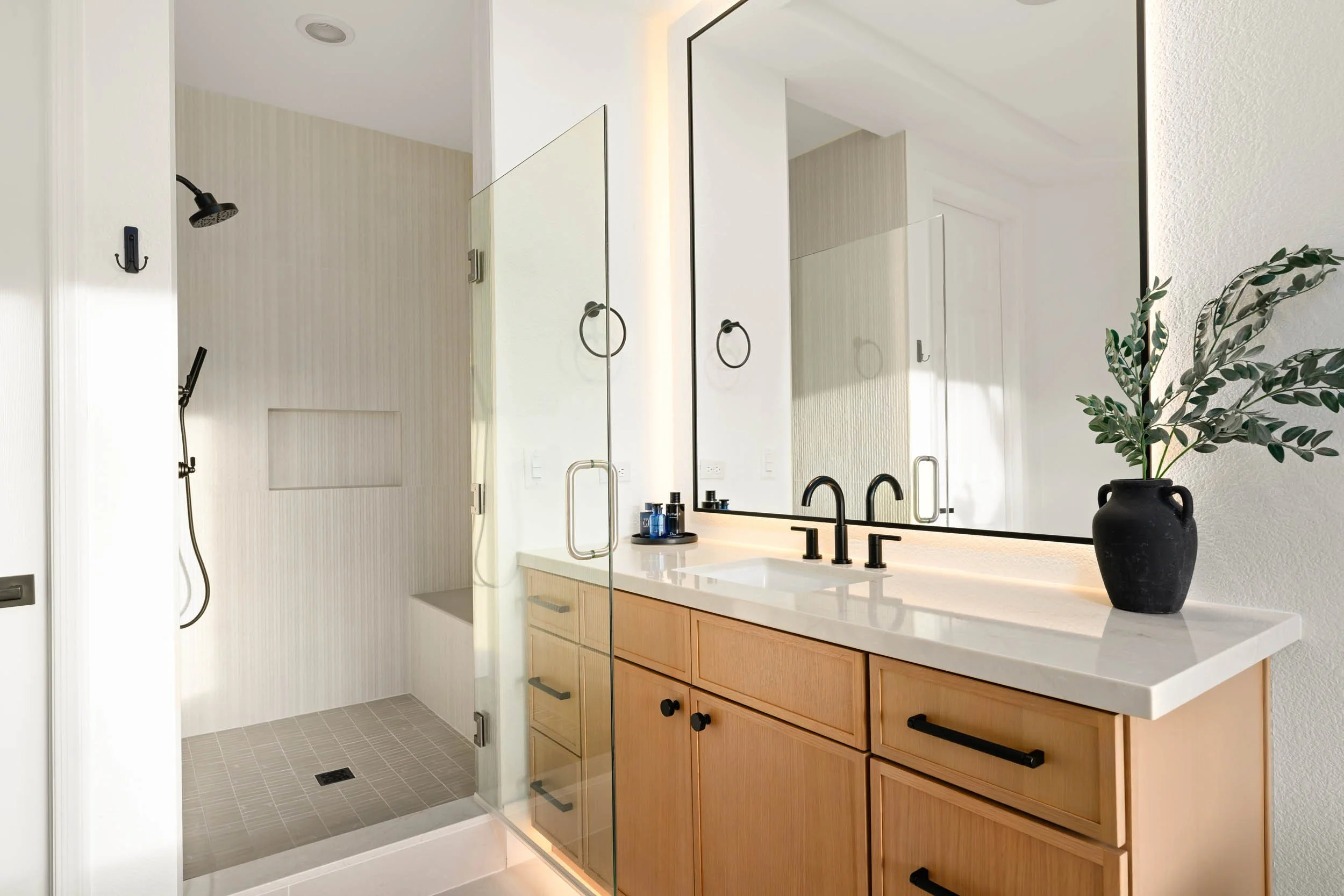 Modern bathroom with a walk-in shower, a large mirror, a white countertop with a black faucet, and a black vase with green leaves.