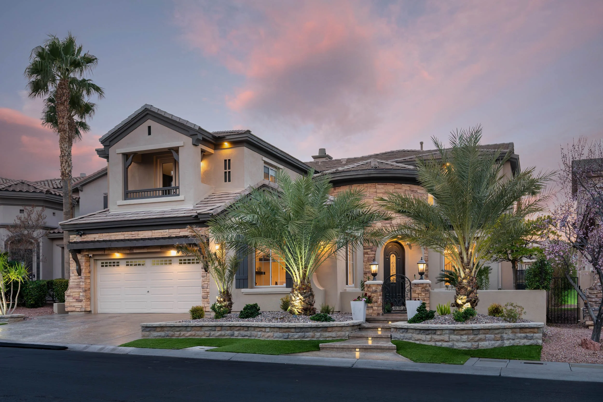 Luxury two-story house with a stone and stucco exterior, tall palm trees, lit front yard, driveway, and a sunset sky in the background.