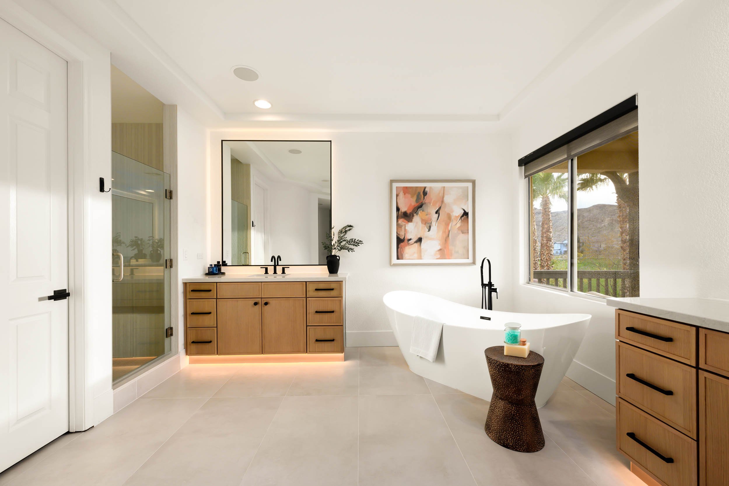 Modern bathroom with white walls, large window, wooden cabinetry, standalone bathtub, framed artwork, potted plant, and natural light.