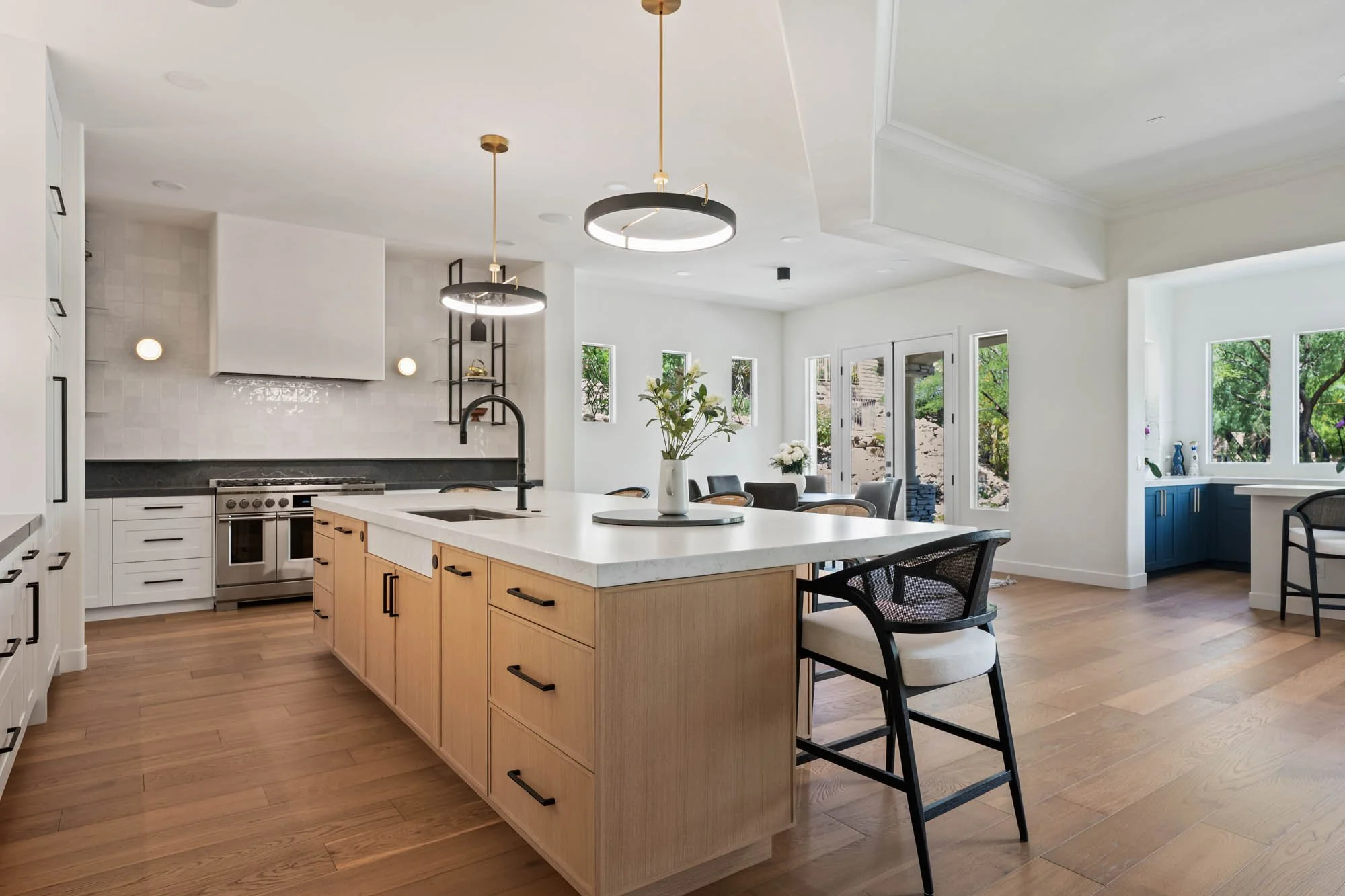 Modern open-concept kitchen with white cabinetry, a large central island with a white countertop, and black hardware. Light wood flooring, black pendant lights, and a vase with green foliage on the island. Walls are white with natural light from mult
