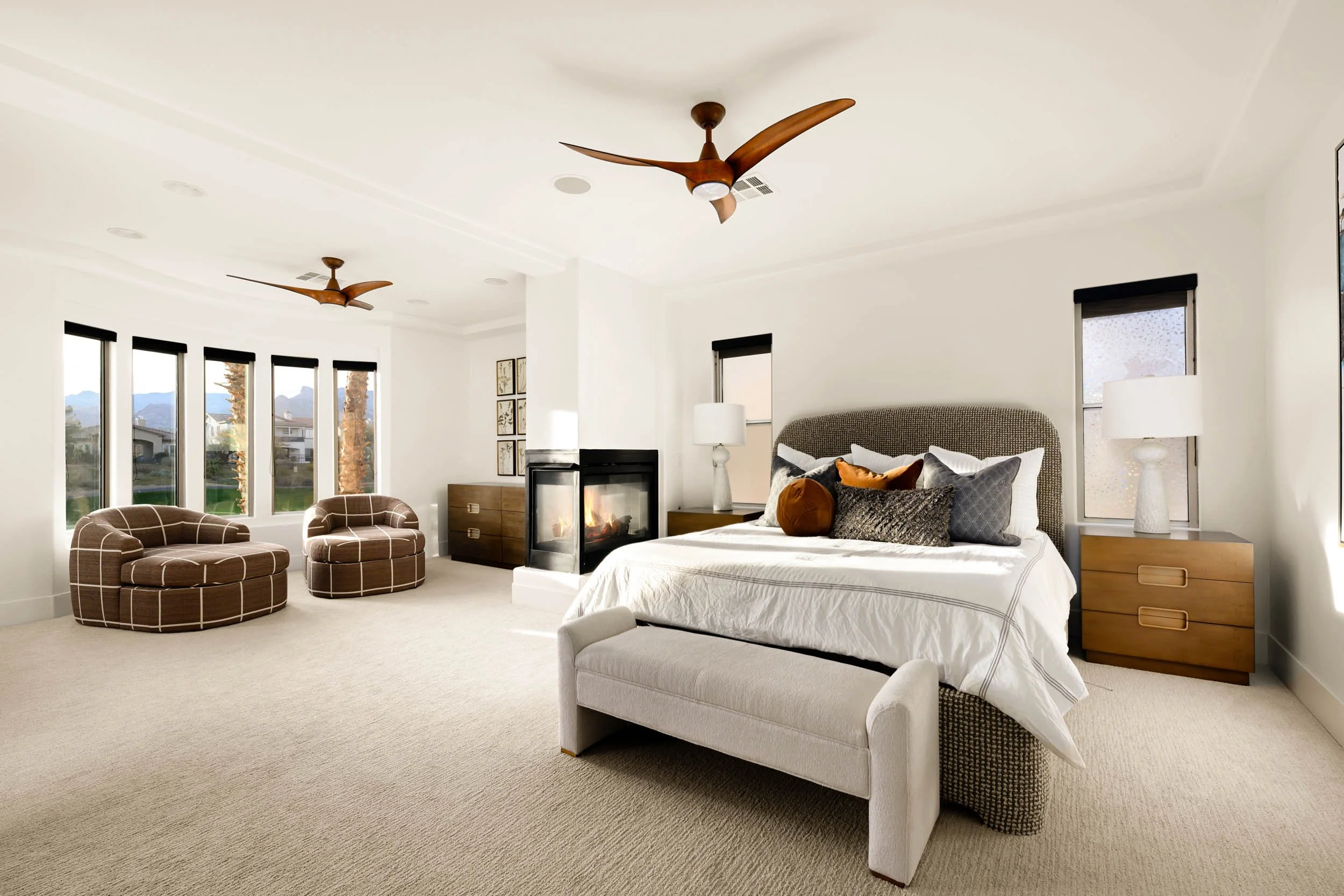 Bright bedroom with a large bed, nightstands with lamps, a fireplace, and sitting area with two brown upholstered chairs near windows showing a scenic outdoor view.