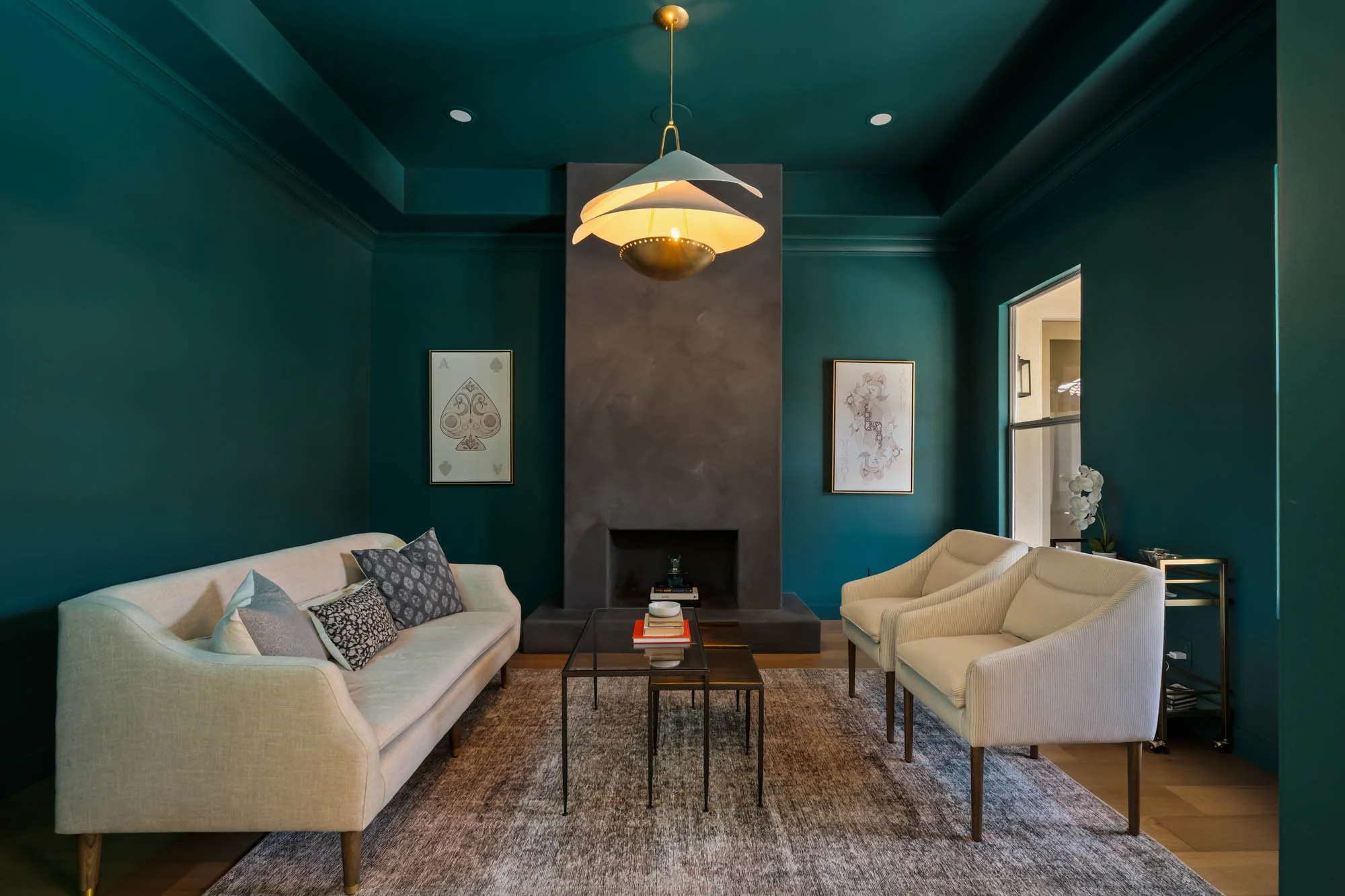 Living room with teal walls, white sofa with patterned pillows, two white armchairs, black coffee table with books, dark gray fireplace, framed artwork, window on right, gold and white ceiling light, beige area rug.