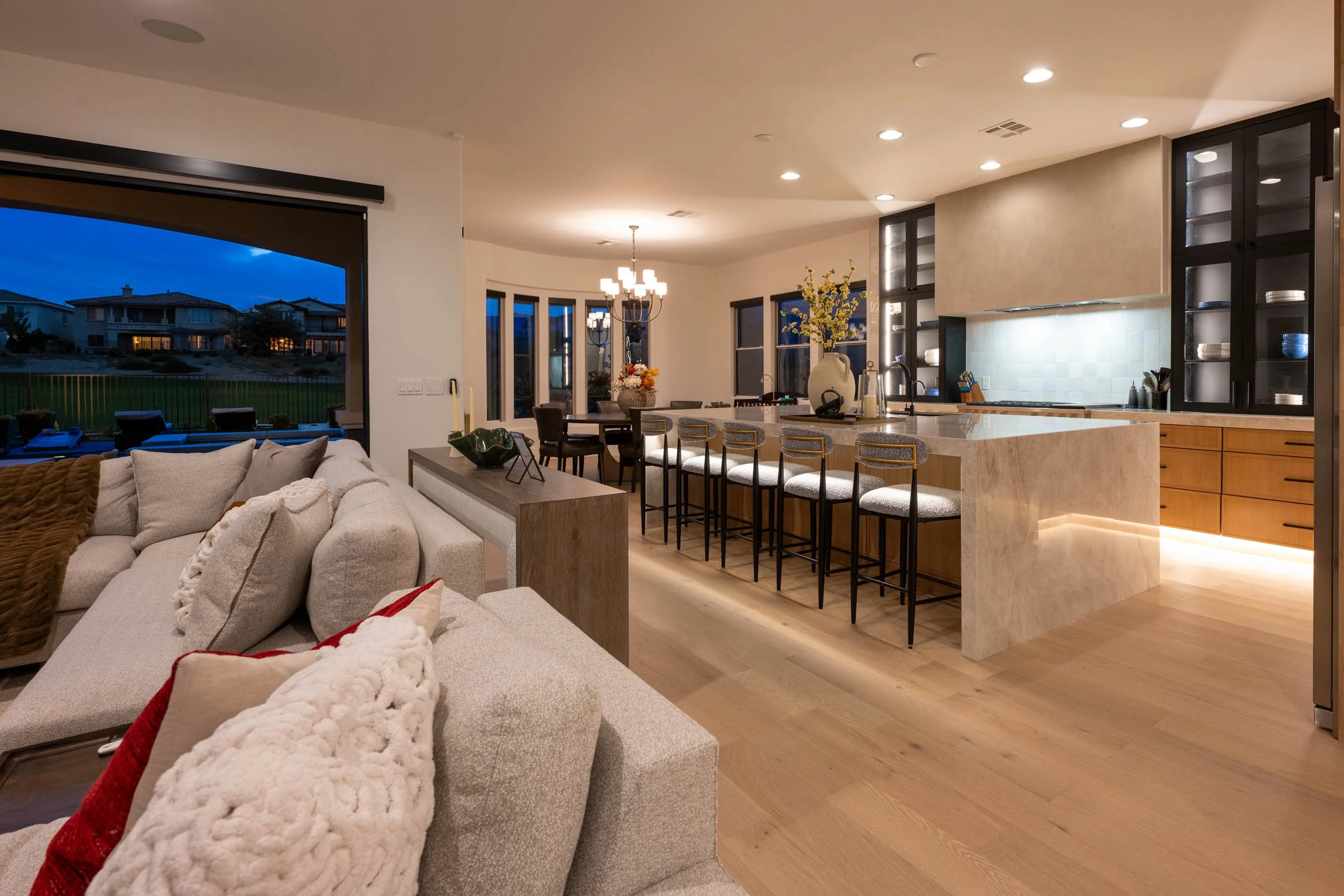 Open-concept living room and kitchen area in a modern home at dusk. The living room has beige sofas with decorative pillows, and large windows showing outdoor houses and a lawn. The kitchen features a marble island with barstools, wooden cabinets, bl