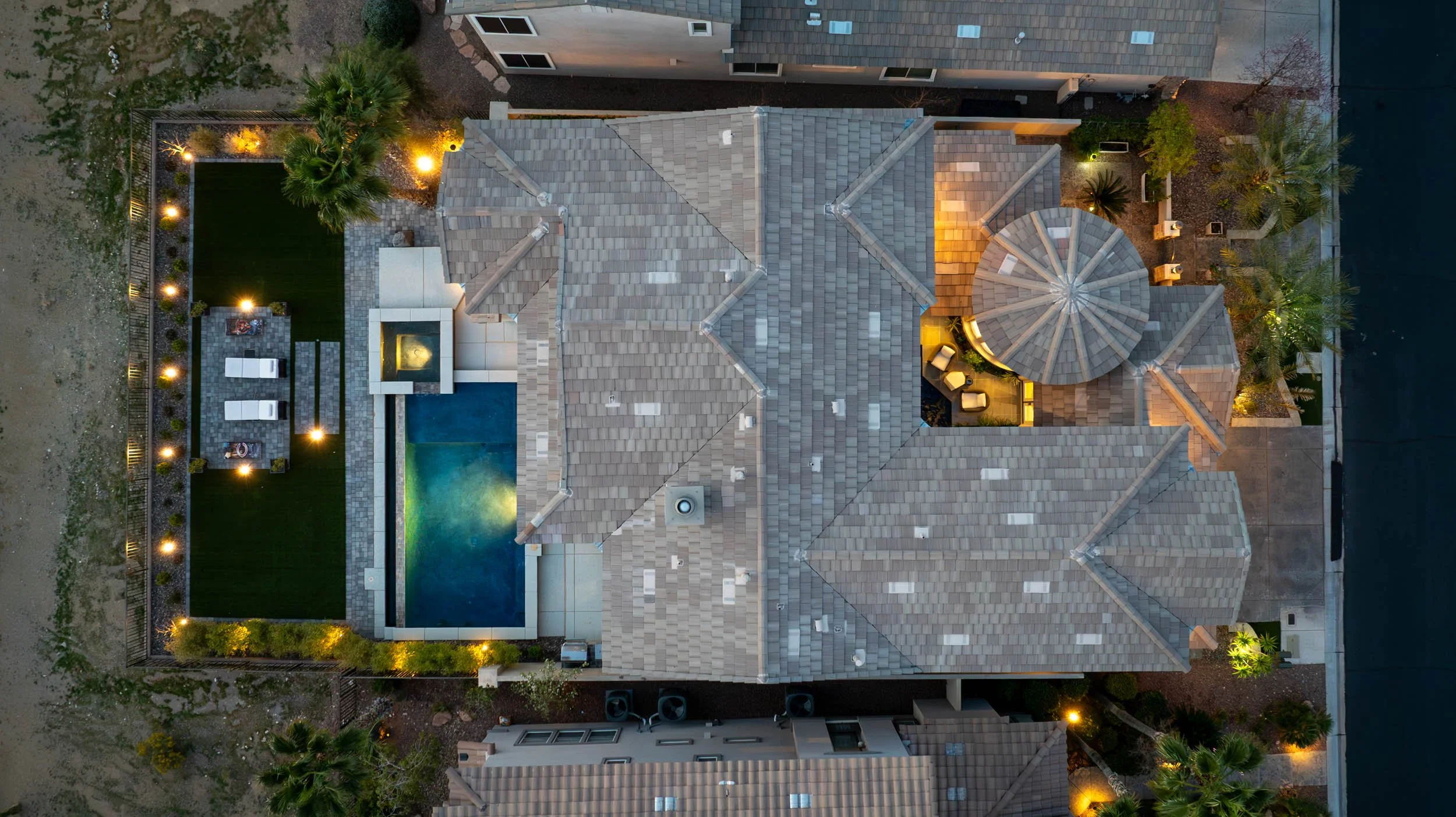An aerial view of a house with a tiled roof, outdoor pool, patio furniture, and landscaped yard at night with outdoor lighting.