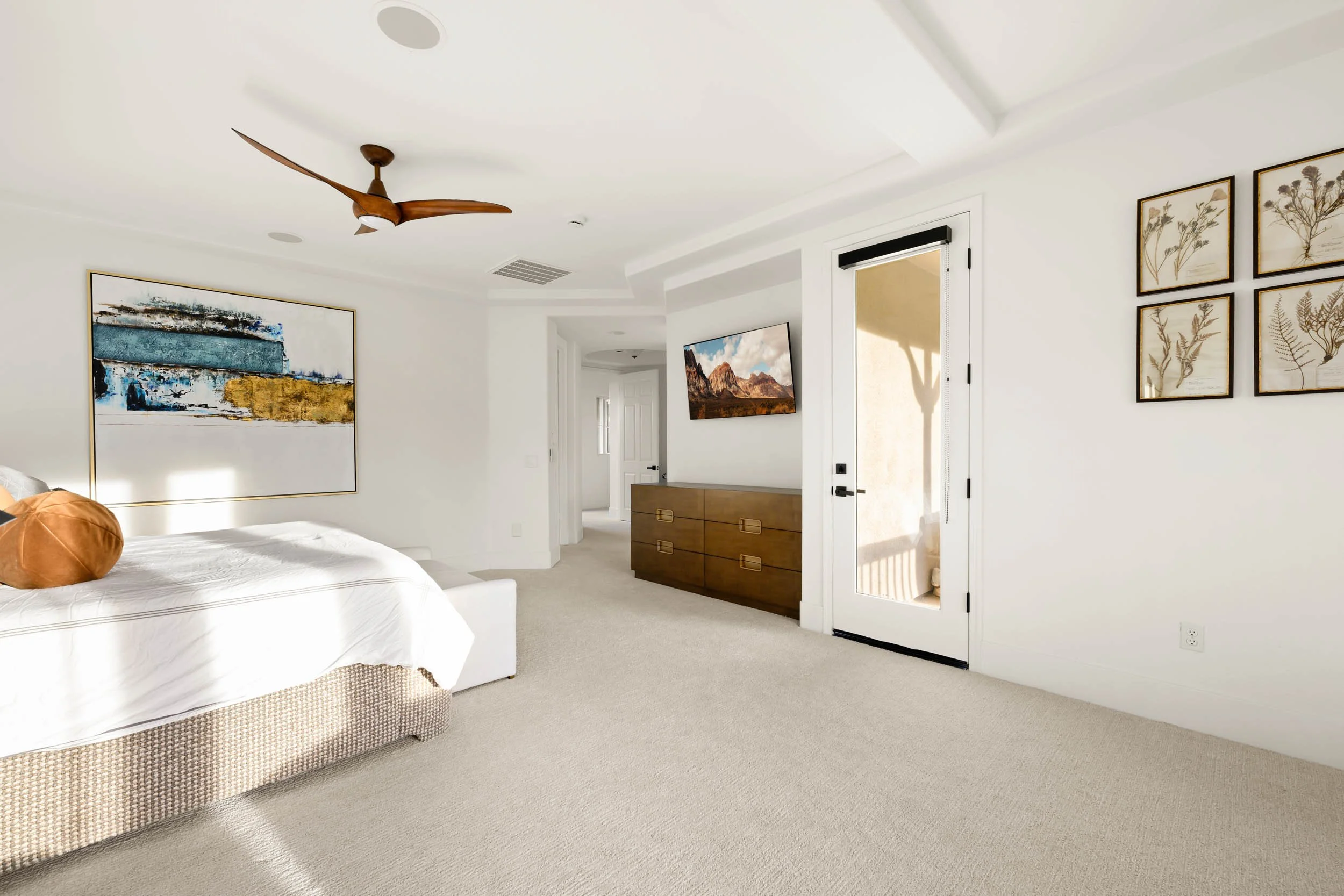 Bright hotel bedroom with white walls, a bed with white bedding, a large blue and gold abstract painting, a wooden dresser, a small wall-mounted television, framed botanical prints, a ceiling fan, and a glass door leading to a balcony.