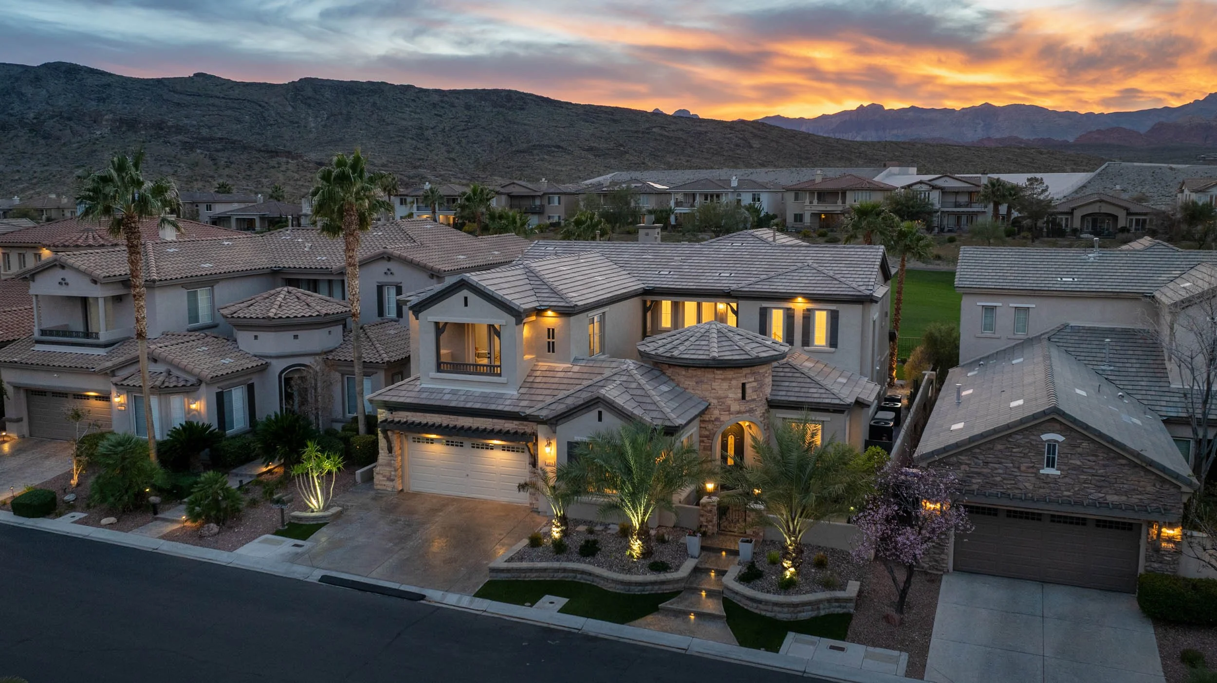 A neighborhood street at sunset showcasing a large modern house with a tiled roof, palm trees, and well-lit exterior, surrounded by similar residential homes, with mountains and a colorful sky in the background.