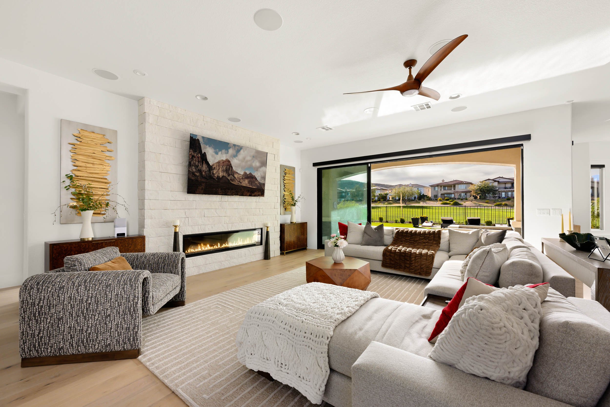 Modern living room with a large L-shaped sofa, armchair, and a glass sliding door overlooking a backyard with grass and houses in the distance. Features include a white brick fireplace, wall-mounted TV, ceiling fan, and decorative wall art.