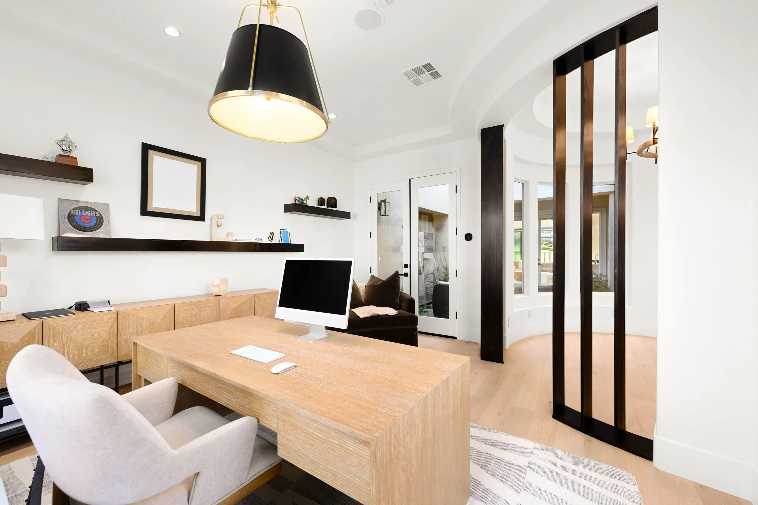 Modern home office with a wooden desk, an iMac computer, a cozy armchair, and decorative black shelves on white walls.
