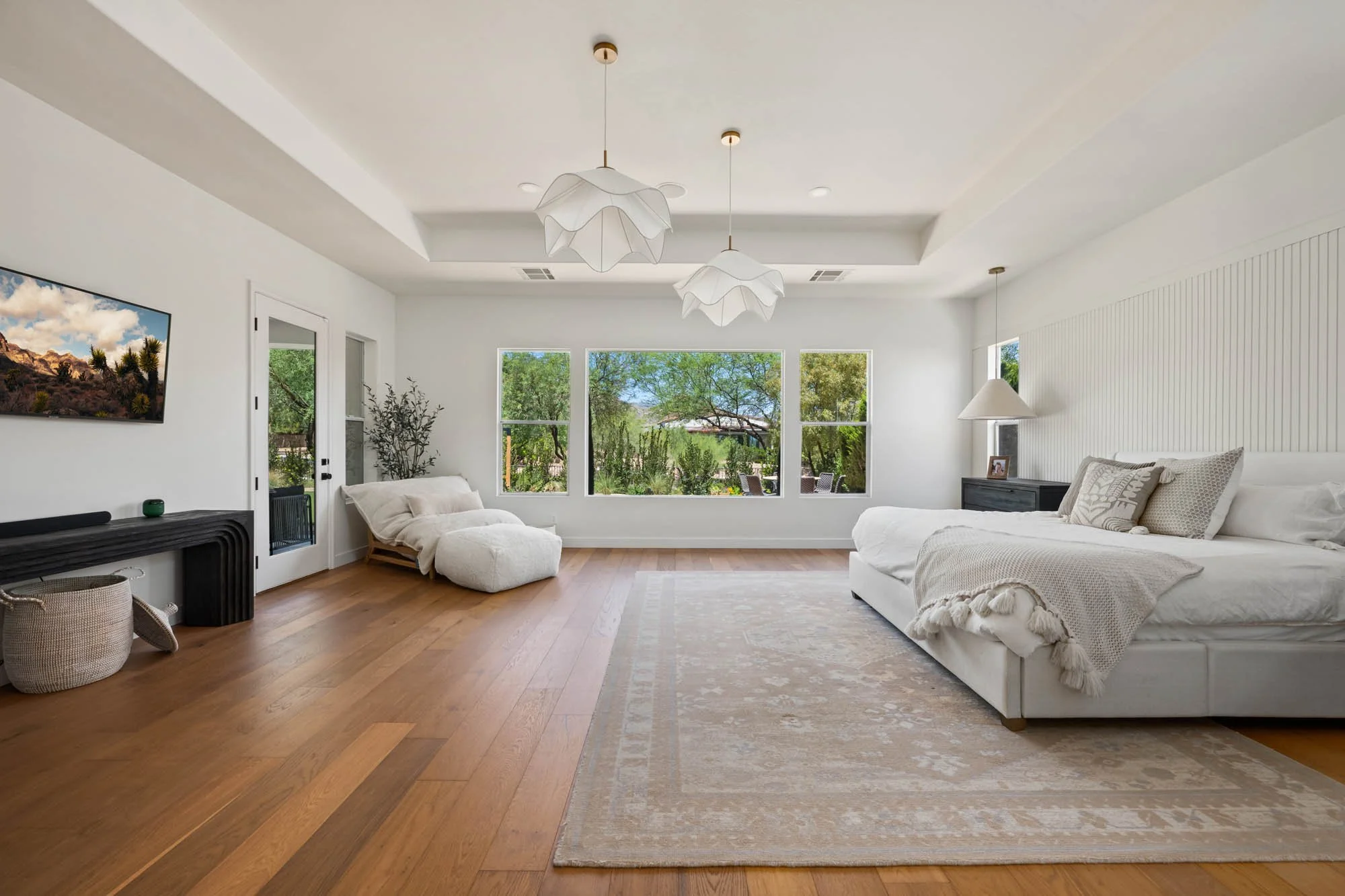 Bright living room with white walls, hardwood floors, large window view of trees, white sofa with pillows, cream blanket, black side table with picture frame, wall-mounted TV, hanging pendant lights, and cozy lounge chair.