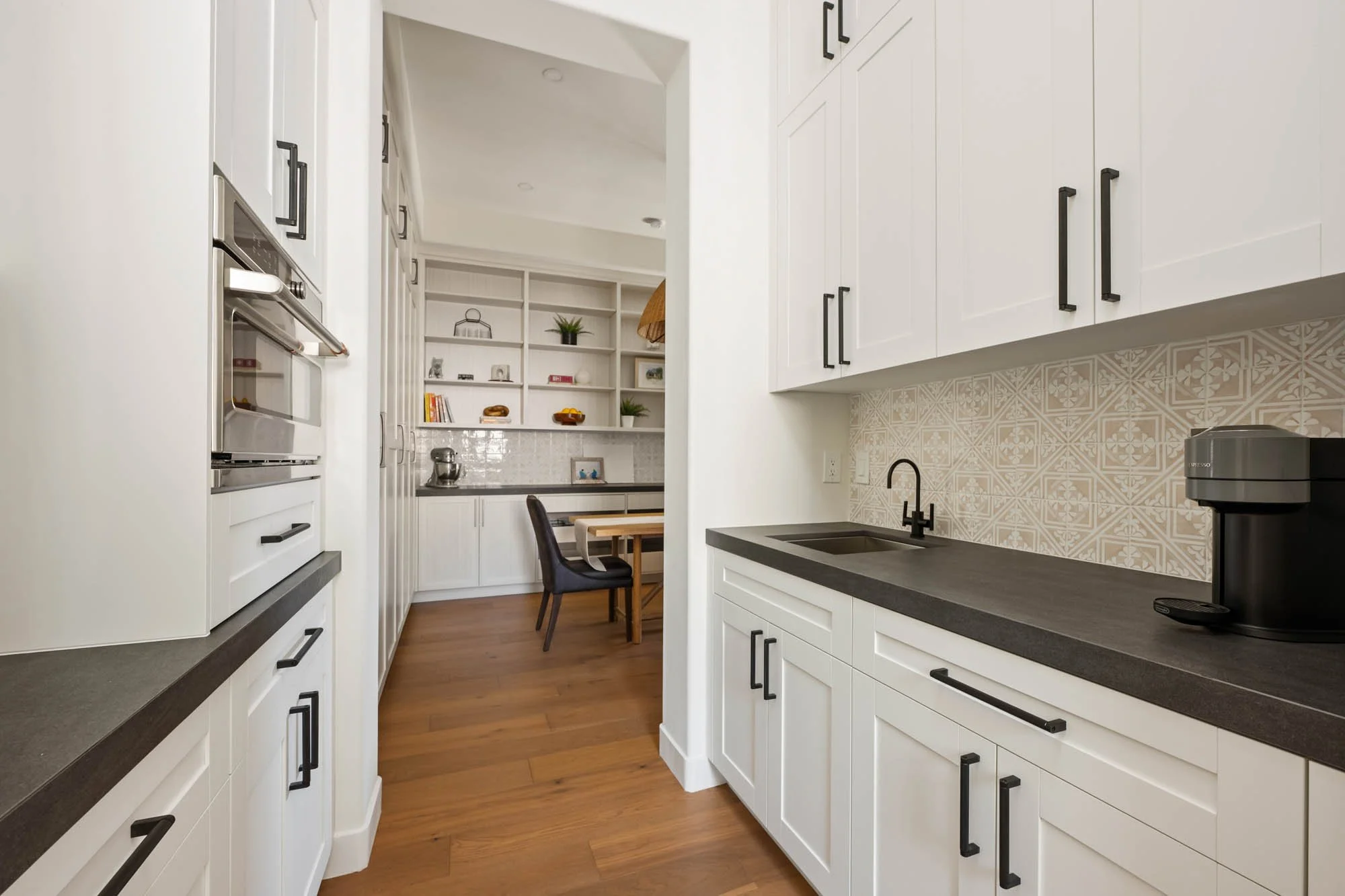 Modern kitchen with white cabinets, black handles, black faucet, black coffee maker, gray countertops, and decorative tile backsplash, leading to a dining area with a table and chairs and built-in shelves.