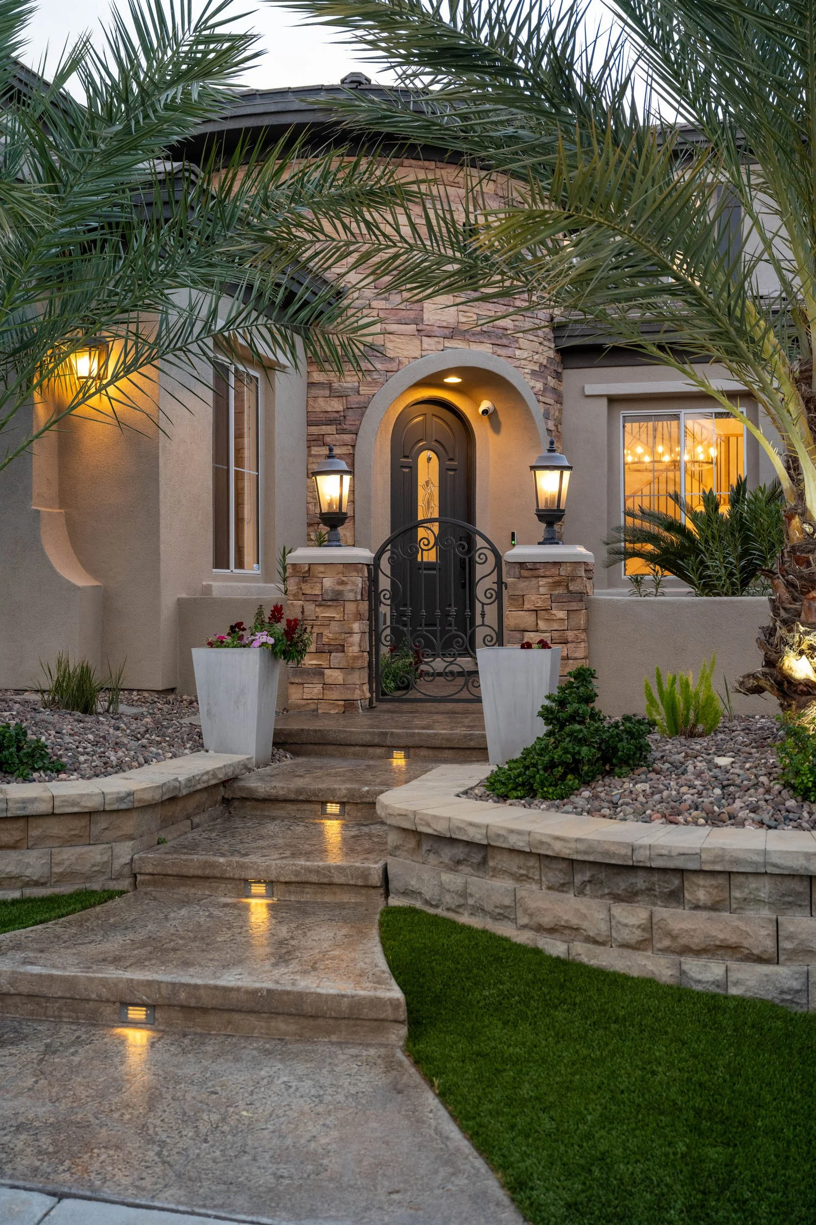 Front entrance of a house with stone steps, a black metal gate, large potted plants, palm trees, and warm exterior lighting