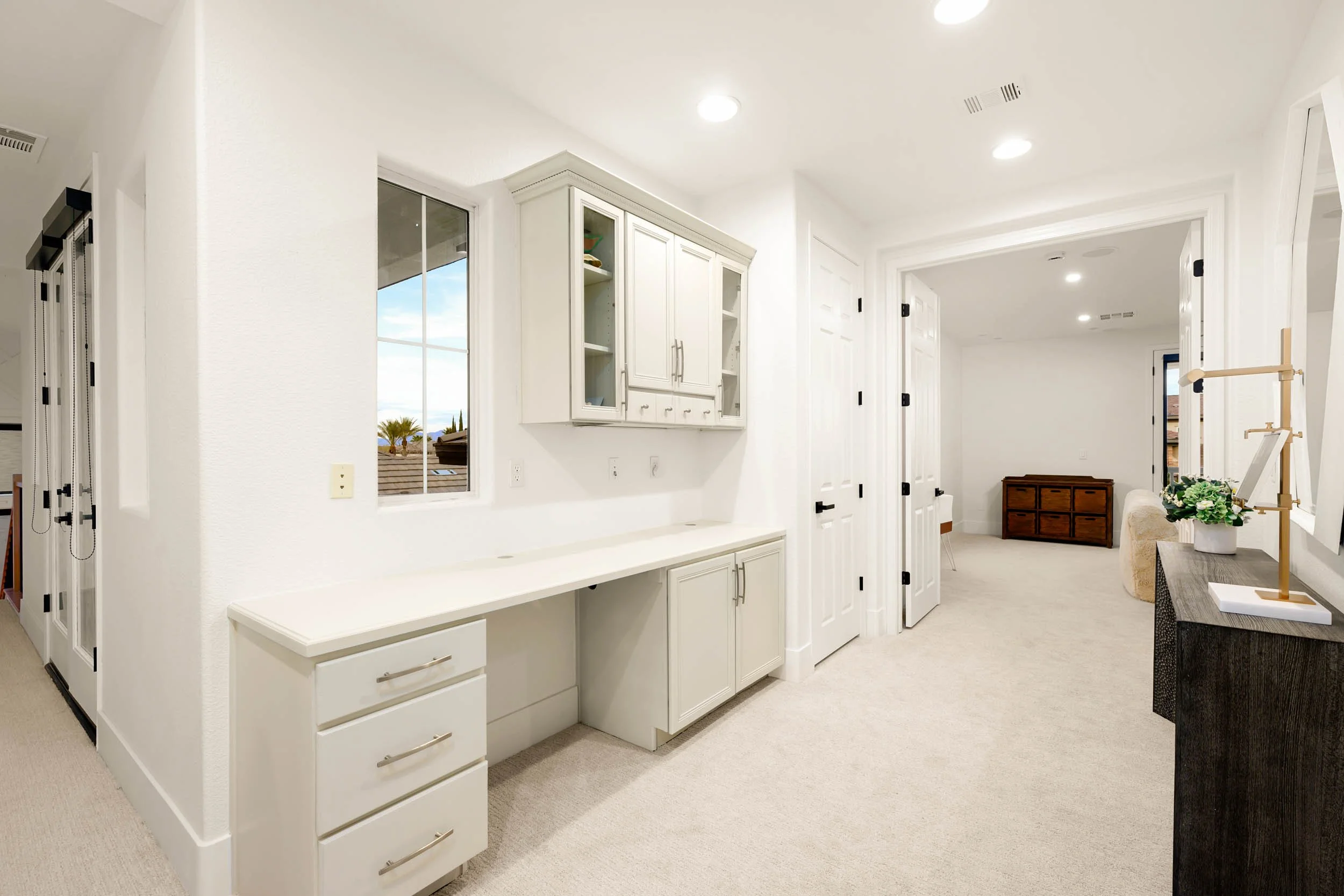 Bright, modern living space with white cabinetry, windows showing an outdoor view, and beige carpeted flooring.
