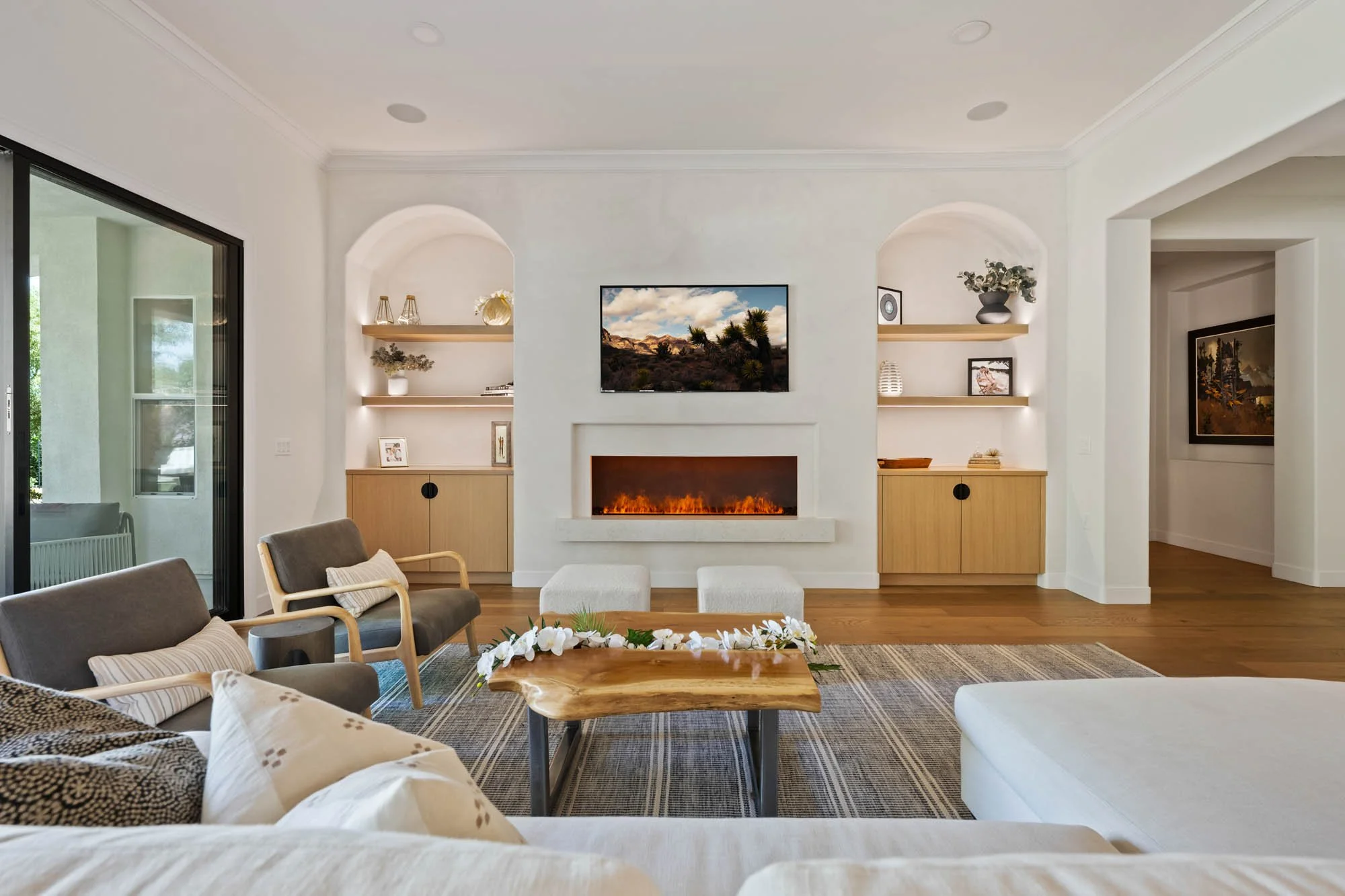 Living room with white walls, wooden floor, and a modern fireplace under a wall-mounted TV. There are armchairs, a coffee table with flowers, built-in shelves with decor, and a sliding glass door leading outside.