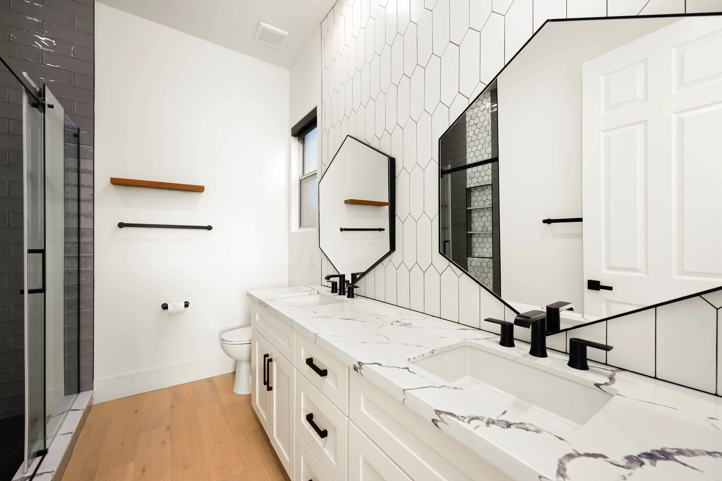 Modern bathroom with white vanity, black hardware, large mirror with geometric black trim, black faucets, black-tiled shower, hexagonal wall tiles, wooden floor, and white walls.