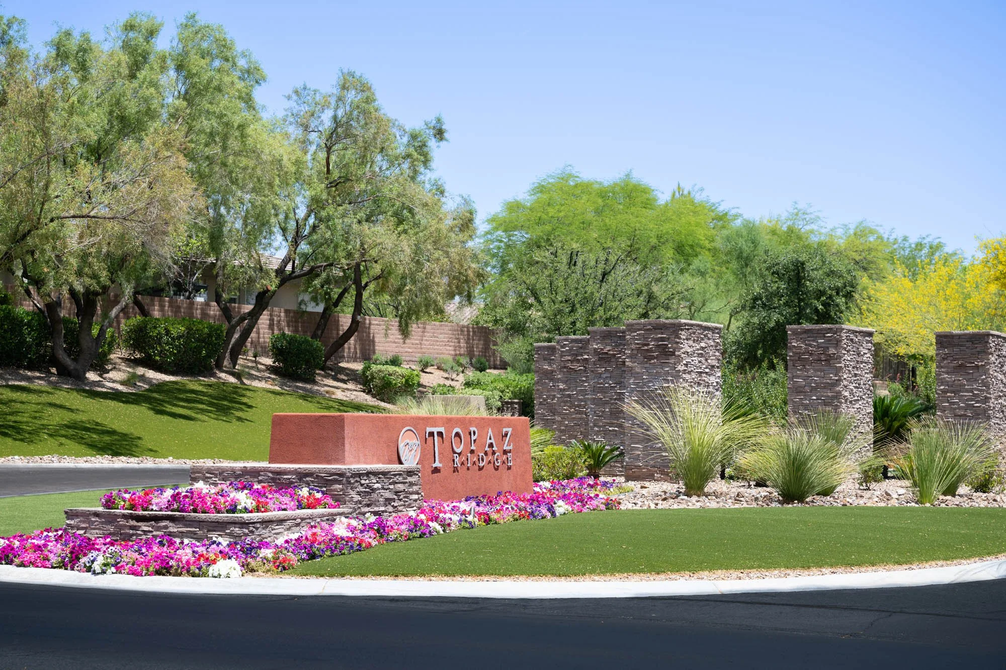 Entrance sign for Topaz Ridge community situated on a landscaped roundabout with colorful flowers, stone pillars, and trees in the background under a clear blue sky.