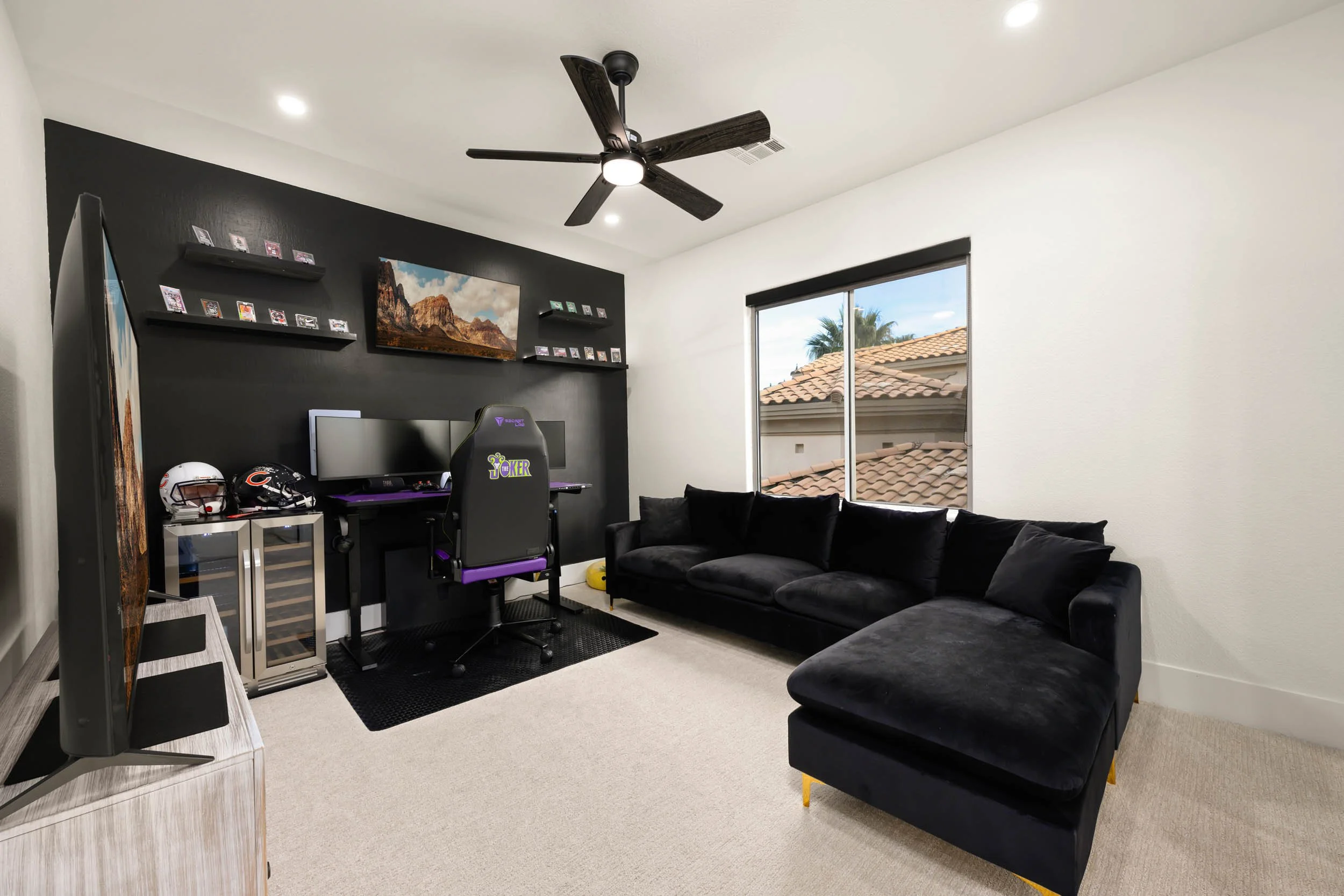 A modern living room with a black and white color scheme, featuring a large black sectional sofa, a window with a view of a rooftop and palm trees, a ceiling fan, a black accent wall with wall-mounted shelves, a computer and gaming setup, and a flat-