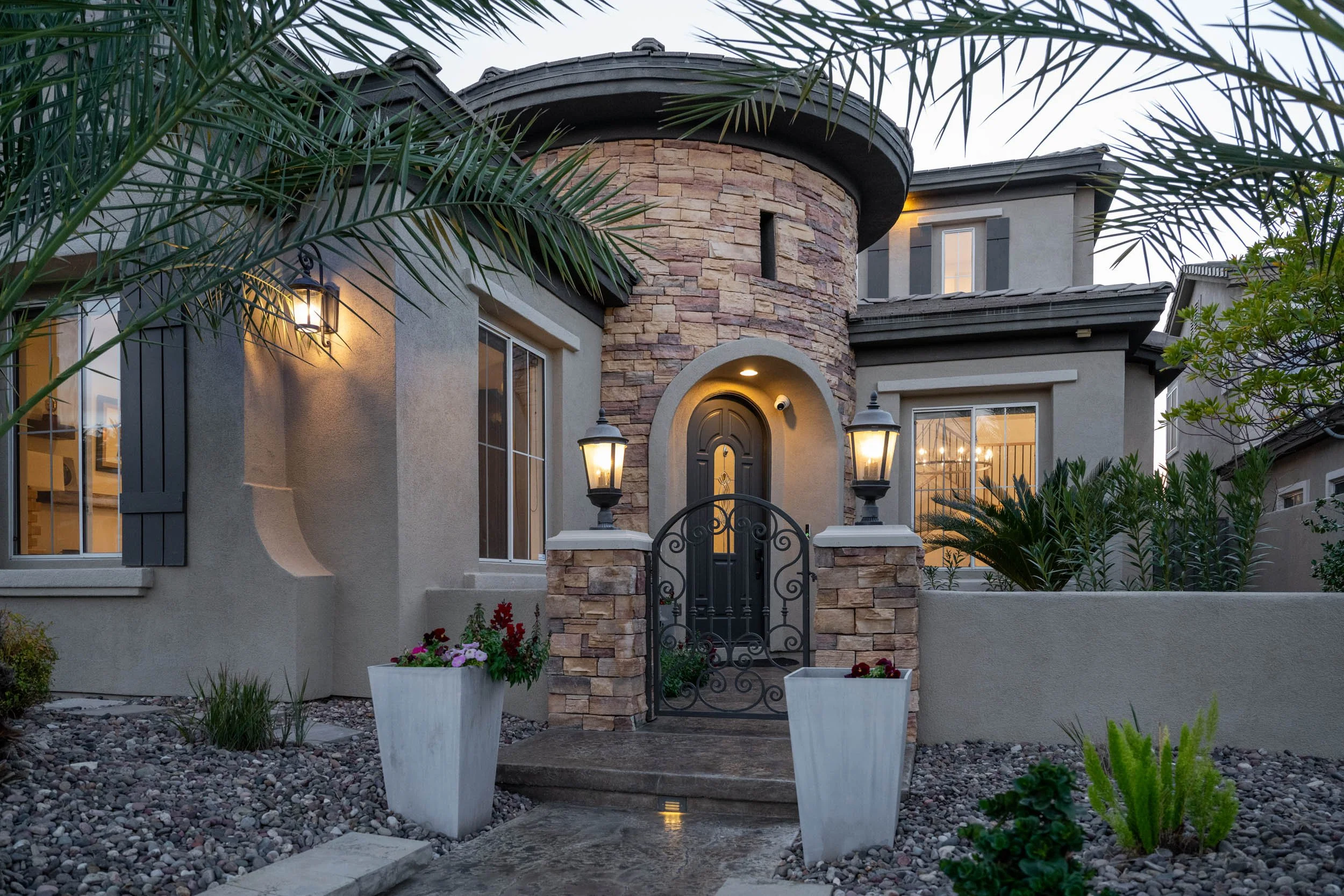 Front view of a modern house with a stone turret, black wrought iron gate, and warm exterior lighting. Smartly landscaped with potted plants and desert-style gravel.