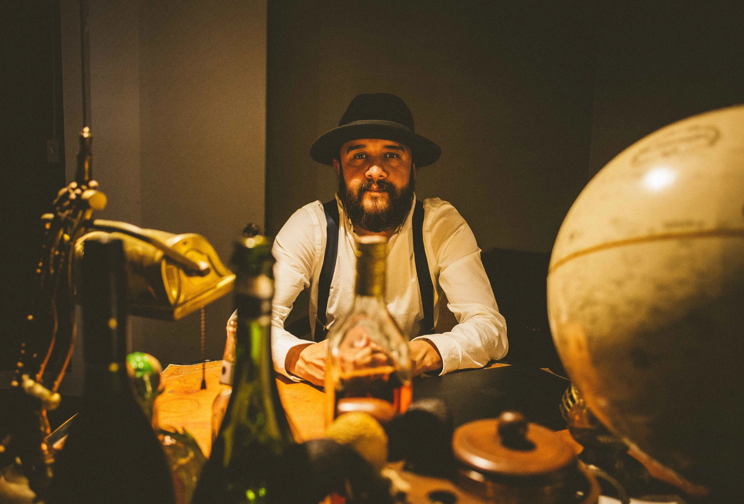 A man with a beard wearing a white shirt, suspenders, and a black hat sits at a cluttered desk with vintage items, including a globe, bottles, and decorative objects, in a dimly lit room.