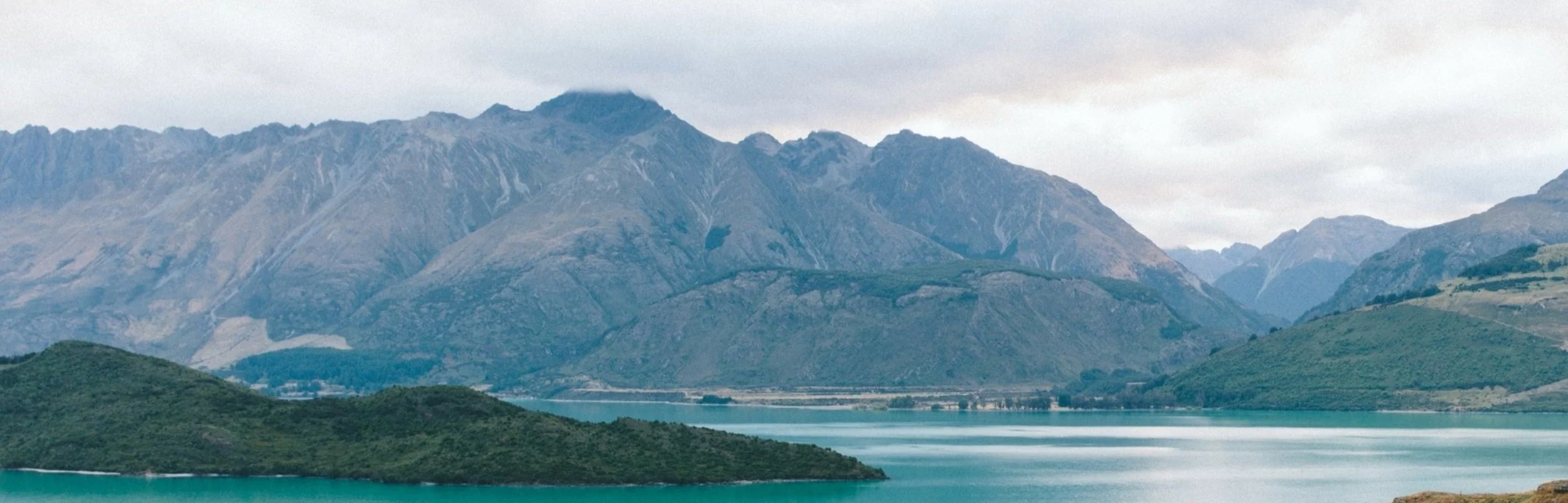 Mountain range with a body of water in the foreground under a cloudy sky.