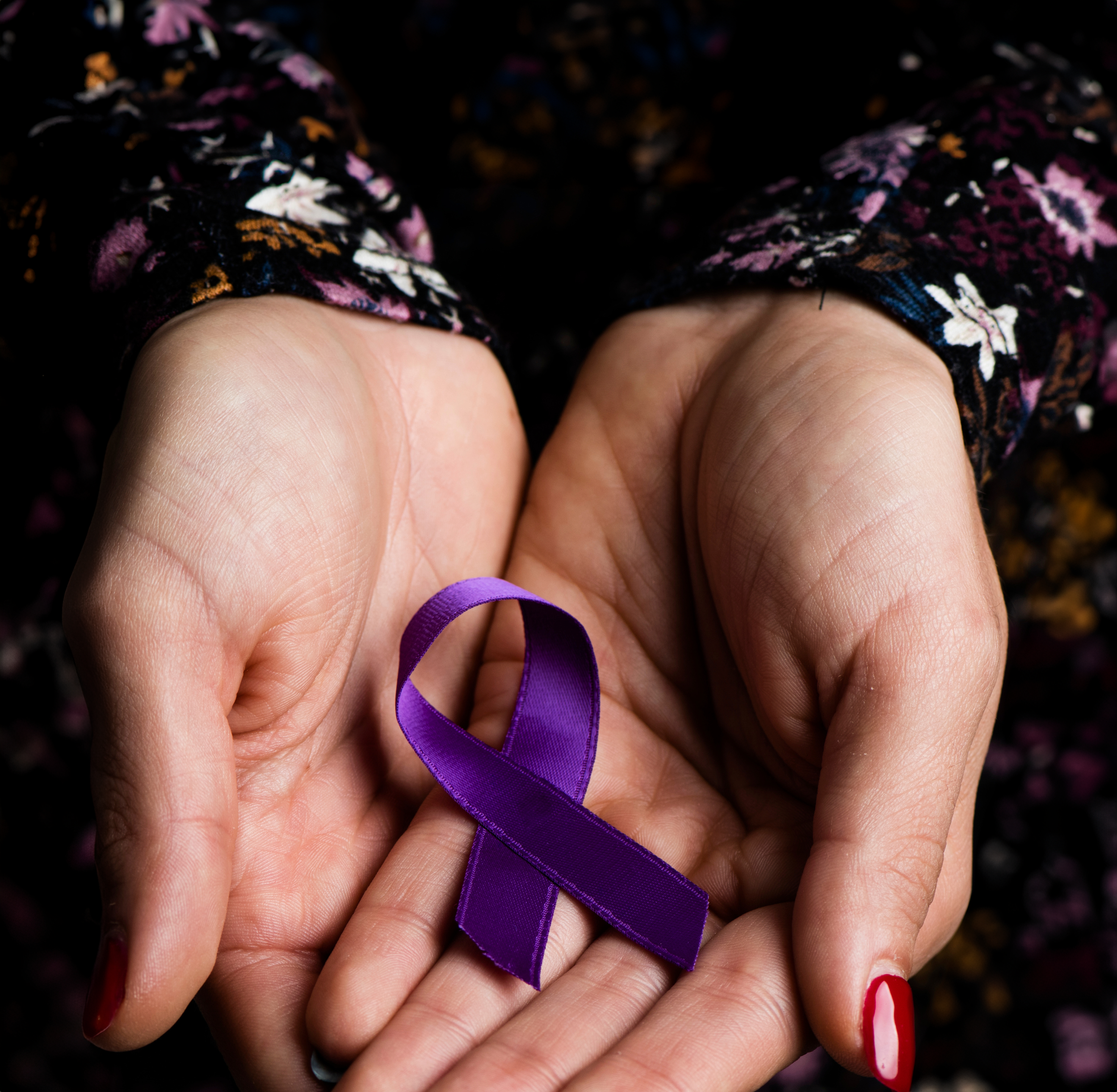 A close-up, eye-level shot of a woman's cupped hands gently holding a vibrant purple awareness ribbon. The overall mood is one of quiet strength and support.