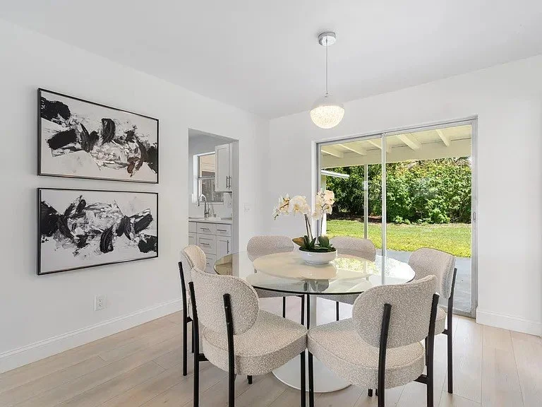 Bright dining room with a round table and six upholstered chairs. A large sliding glass door leads to a backyard with trees. Artwork with abstract black and white brushstrokes decorates the white wall. A ceiling light hangs over the table, and a kitc