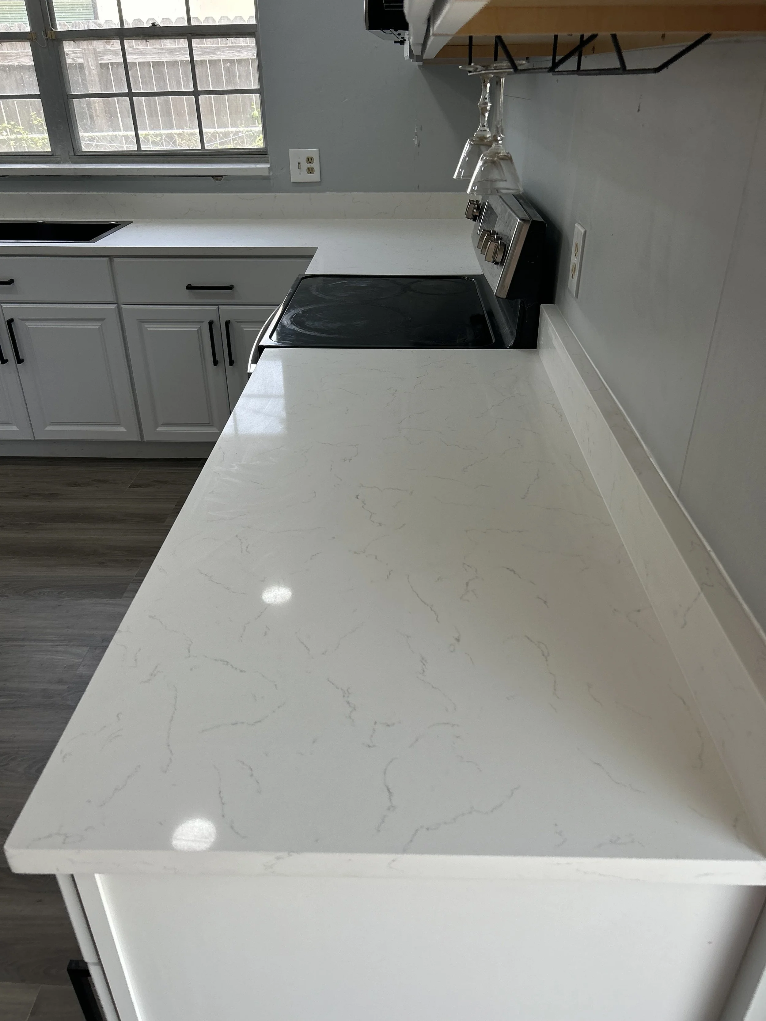 A modern kitchen with white marble countertop, gray cabinets with black handles, stainless steel stove, and a window with blinds.