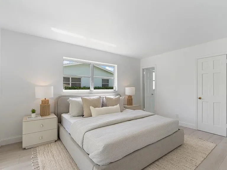 Minimalist bedroom with white walls, a large bed with multiple pillows, two nightstands with lamps, a small plant, and a window showing neighboring houses.