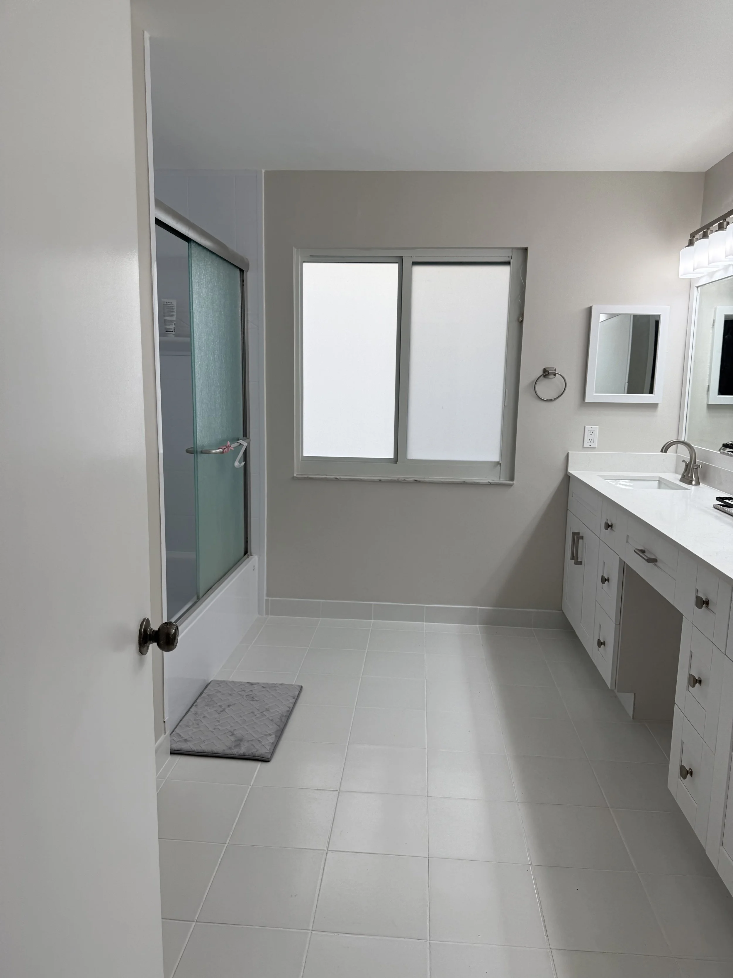 Empty white-tiled bathroom featuring a walk-in shower with sliding glass doors, a window, a white vanity with a sink, a mirror, and a light fixture above. 