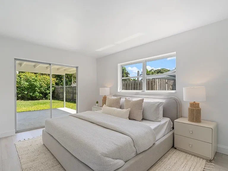 Bright bedroom with white walls, a bed with white bedding and pillows, two white nightstands with lamps, a sliding glass door leading to a green yard, and a window offering a view of a fenced yard and trees.