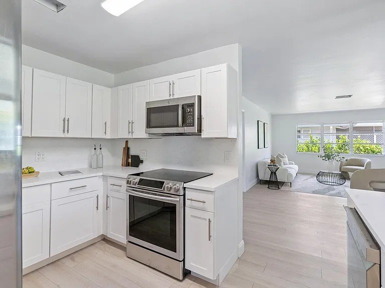 Modern kitchen with white cabinets, stainless steel oven and microwave, light wood flooring, and an open living room with large windows, white sofas, and a black coffee table.