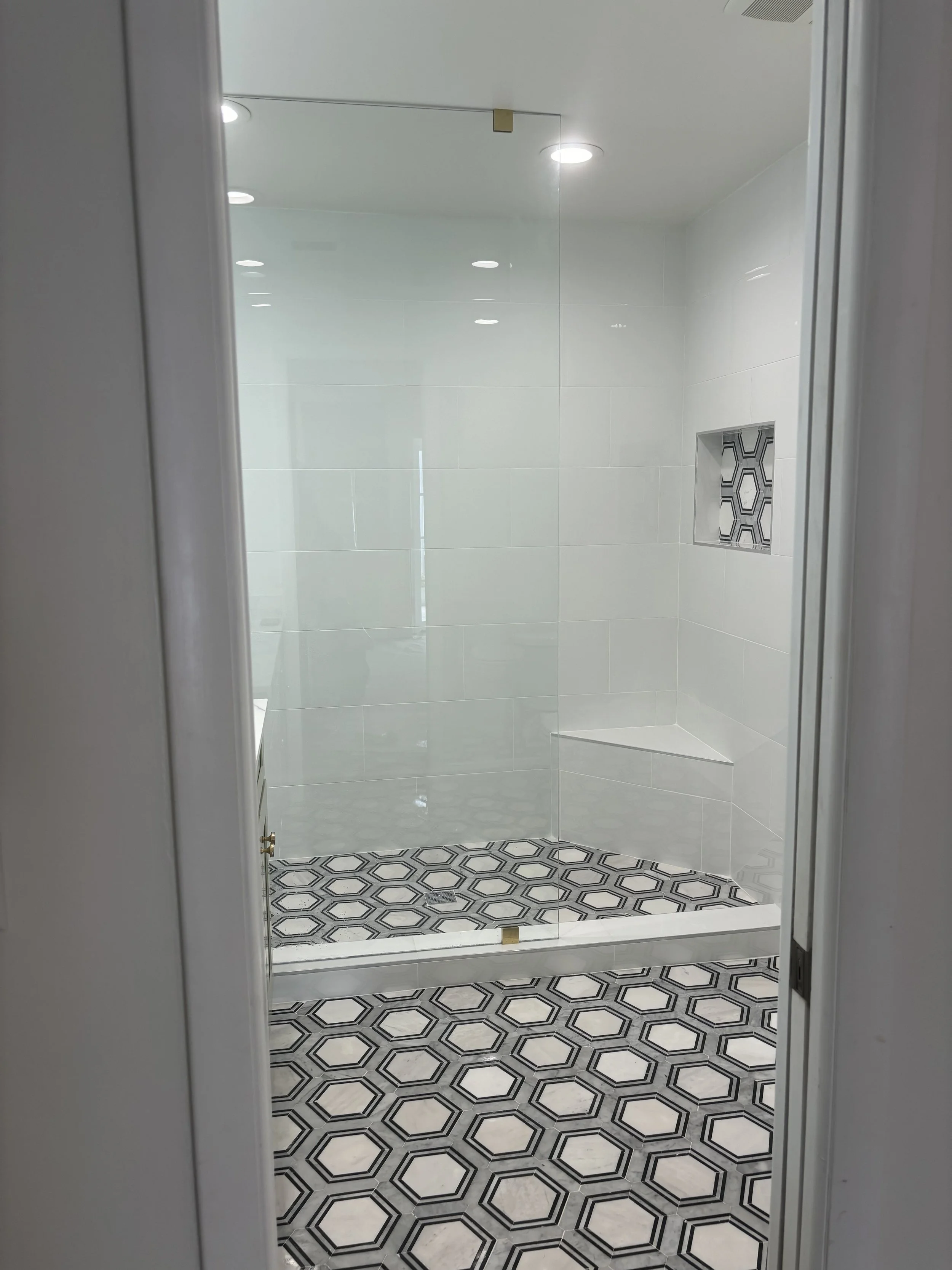 Modern walk-in shower with white tiled walls, a decorative tile niche, and geometric black-and-white patterned floor tiles, seen through a glass door.