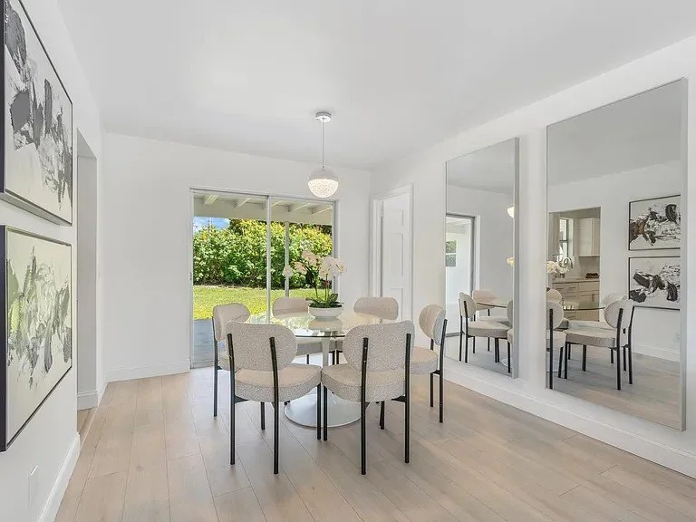 Modern dining room with a round table, seven upholstered chairs, large mirrors on the wall, artwork, a sliding glass door leading to a backyard with greenery, and a view of the kitchen.