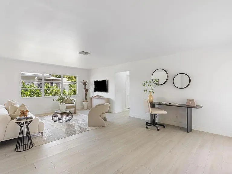 Bright and minimalist living room with white walls, light wood flooring, a white sofa, a black wire coffee table, a flat-screen TV, and a workspace with a desk, chair, and round mirrors on the wall.