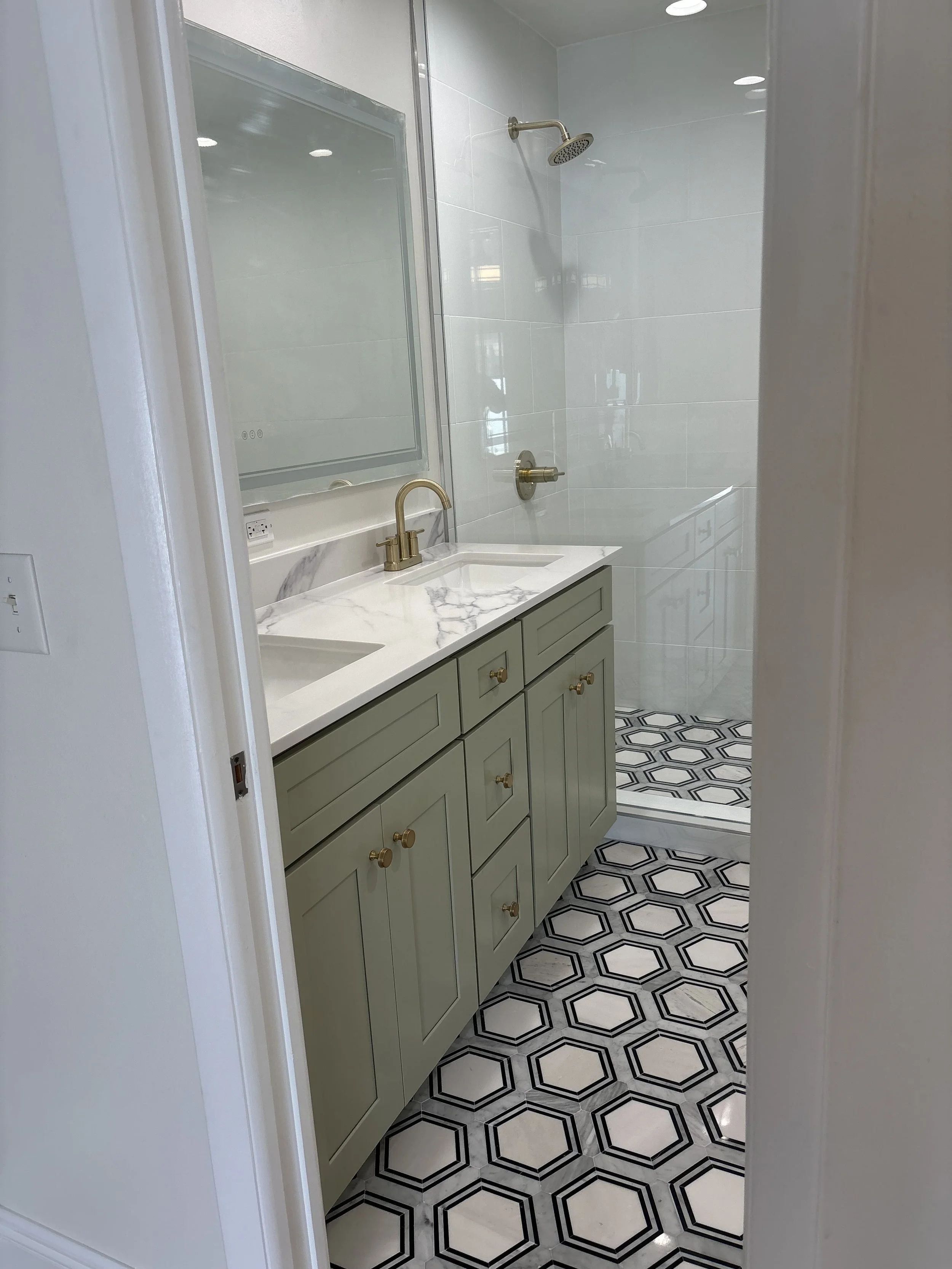 Bathroom with a new double-sink vanity with a Quartz countertop, brass faucet, and a mirror. In the background, there's a walk-in shower with white tiles, a brass showerhead, and patterned black and white hexagonal marble floor tiles.