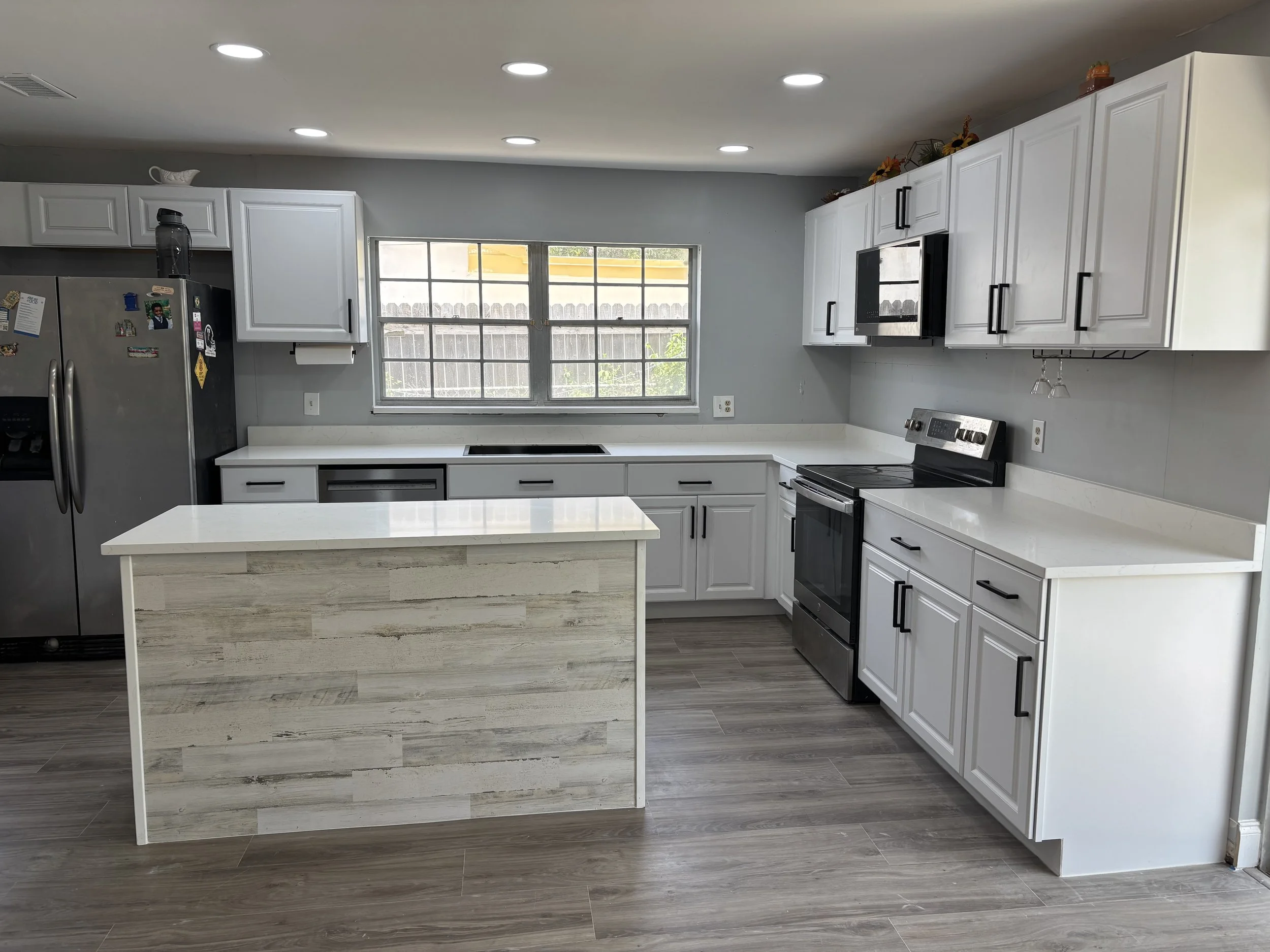 Modern kitchen with white cabinets, black appliances, and a kitchen island with a wood-textured finish, featuring a window over the countertop.