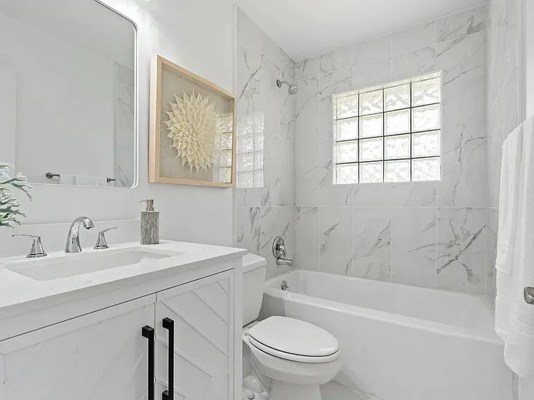 Bright bathroom with white marble walls, a bathtub, a toilet, and a sink with a mirror, decorated with a framed artwork and a small plant.