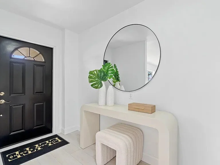 A white foyer table with a round mirror above, two white vases with green monstera leaves, a wooden box, and a plush beige stool underneath, next to a black front door with a welcome mat.