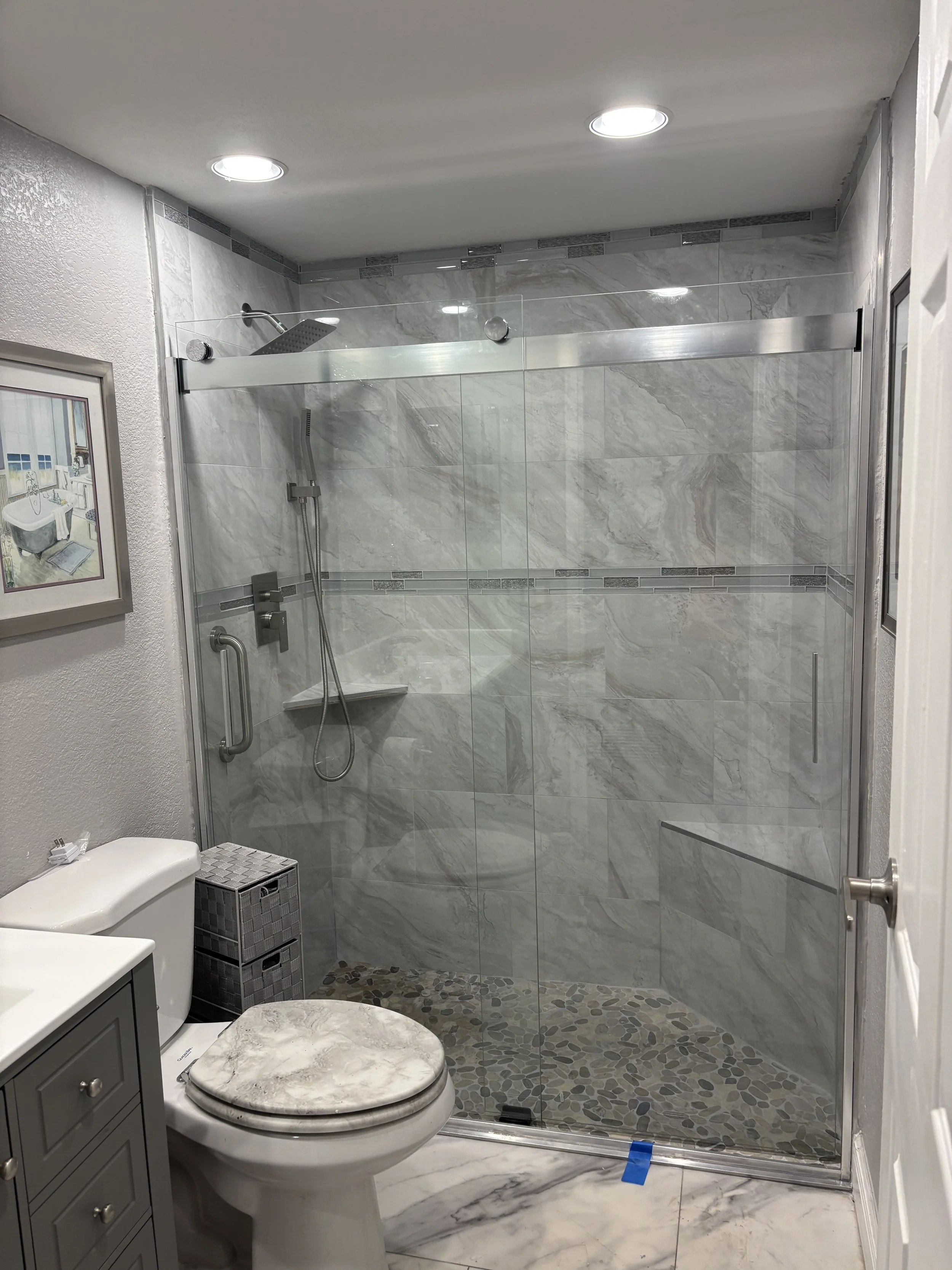 A bathroom with a glass shower enclosure, ceramic tiles, a gray vanity, and a toilet with a marble lid.