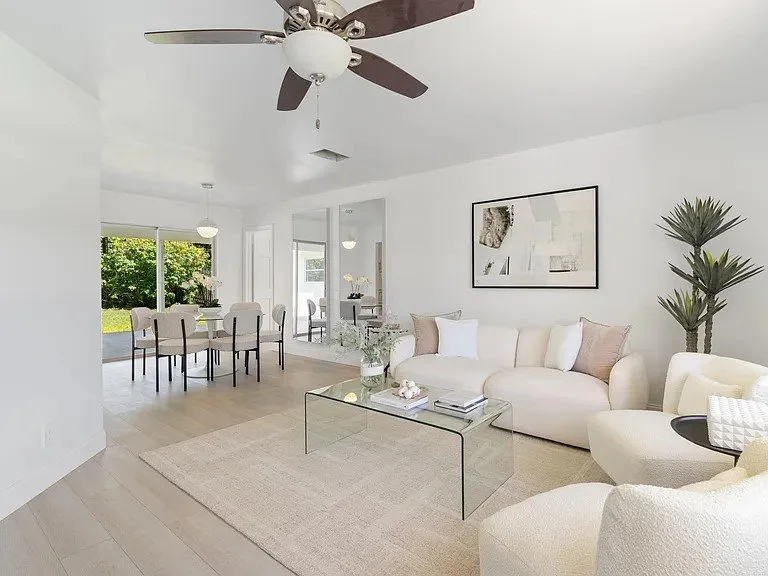 Living room with a white sofa, armchair, glass coffee table, ceiling fan, and wall art. Adjacent dining area with a table and chairs, large sliding door opening to outdoor greenery.