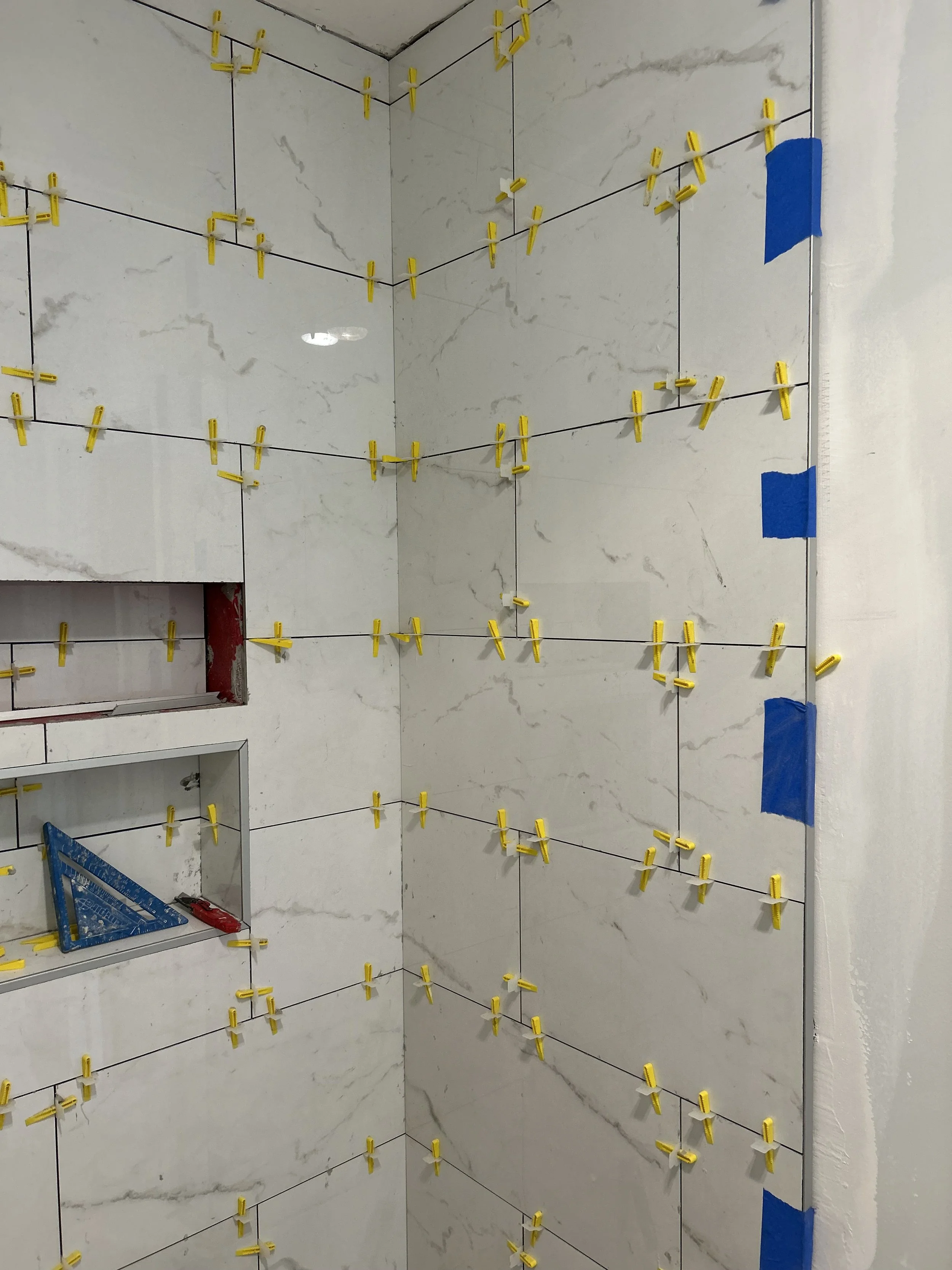 Tile wall with yellow spacers and blue painter’s tape, indicating ongoing tile installation process.