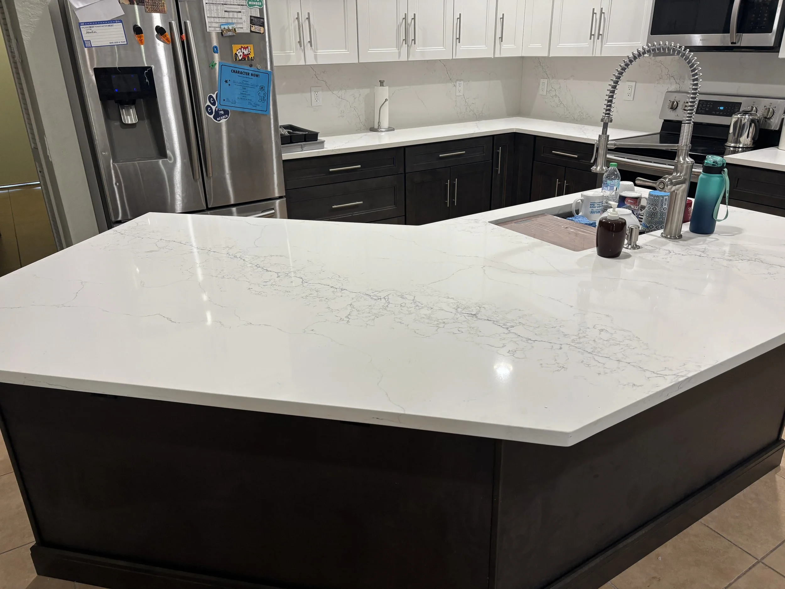 Modern kitchen with white Calcatta  countertop island, dark lower cabinets, white upper cabinets, stainless steel refrigerator, microwave, and oven. Apartment sink with a gooseneck faucet, various kitchen items on the island, and a tiled floor.