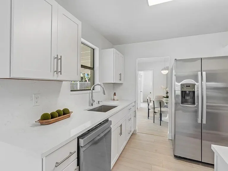 Modern kitchen with white shaker cabinets and Quartz Countertop, a stainless steel refrigerator, a built-in dishwasher, a single-basin sink, and a window above the sink. Light-colored flooring and minimal decor.