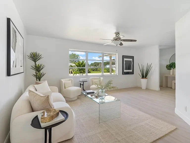 Bright living room with white furniture, large window showing greenery outside, ceiling fan, potted plants, and minimalistic decor.