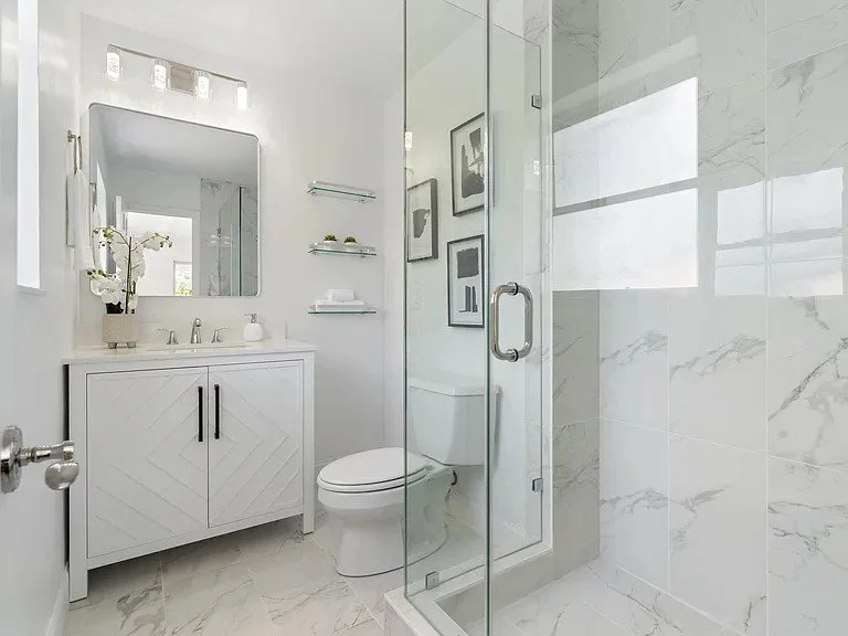 Modern white bathroom with a glass shower enclosure, a white vanity with a mirror, and framed artwork on the wall.