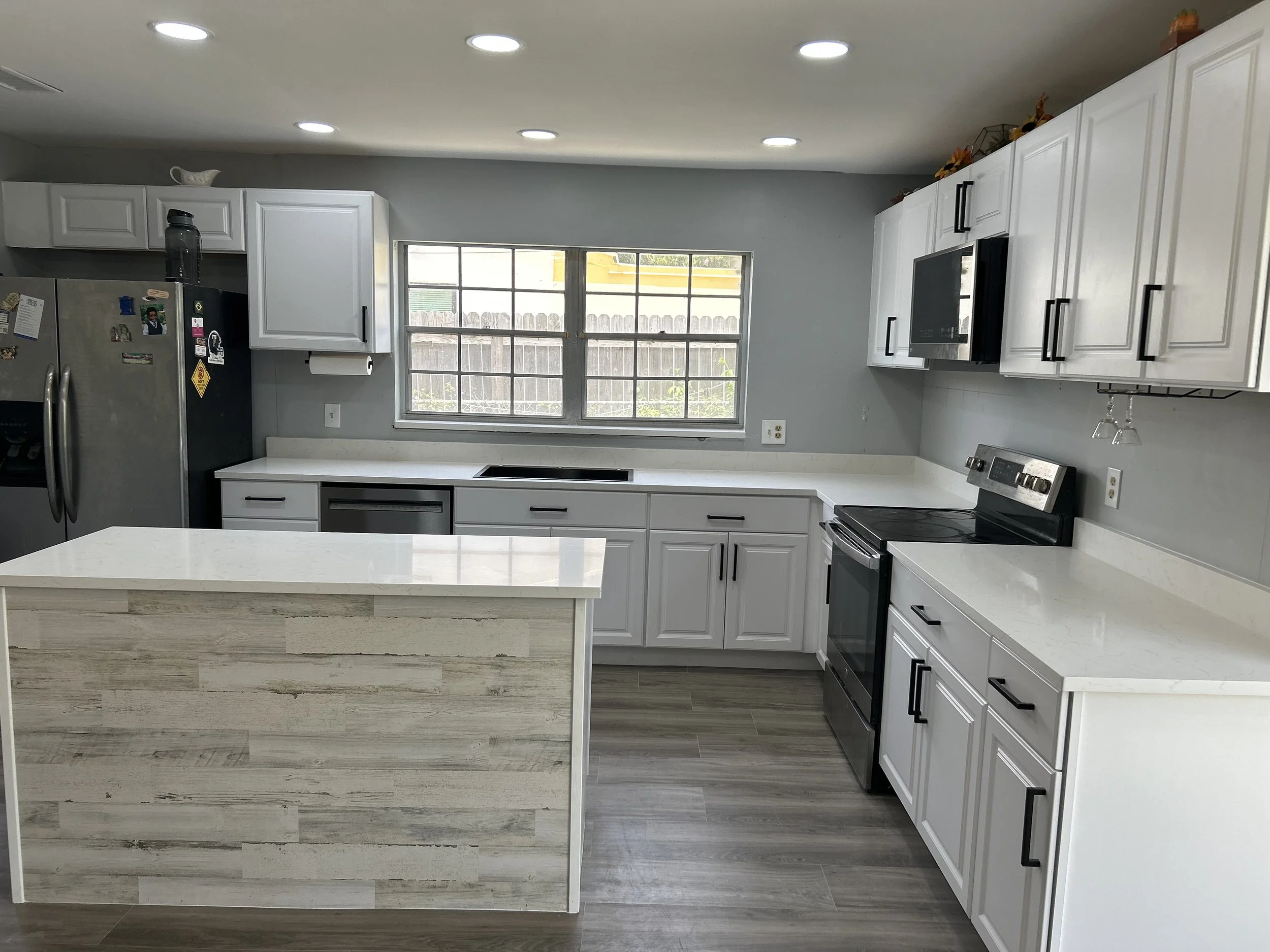 Modern kitchen with white cabinets, black hardware, stainless steel appliances, a central island with a light wood finish, and a large window above the countertop.