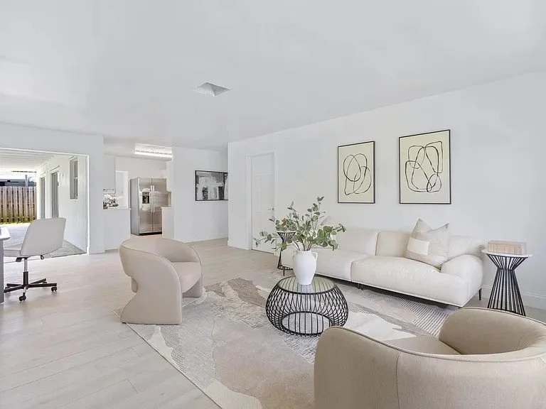Bright living room with white walls, a white sofa, two beige armchairs, and a glass coffee table with a white vase and greenery, with framed abstract art on the wall.